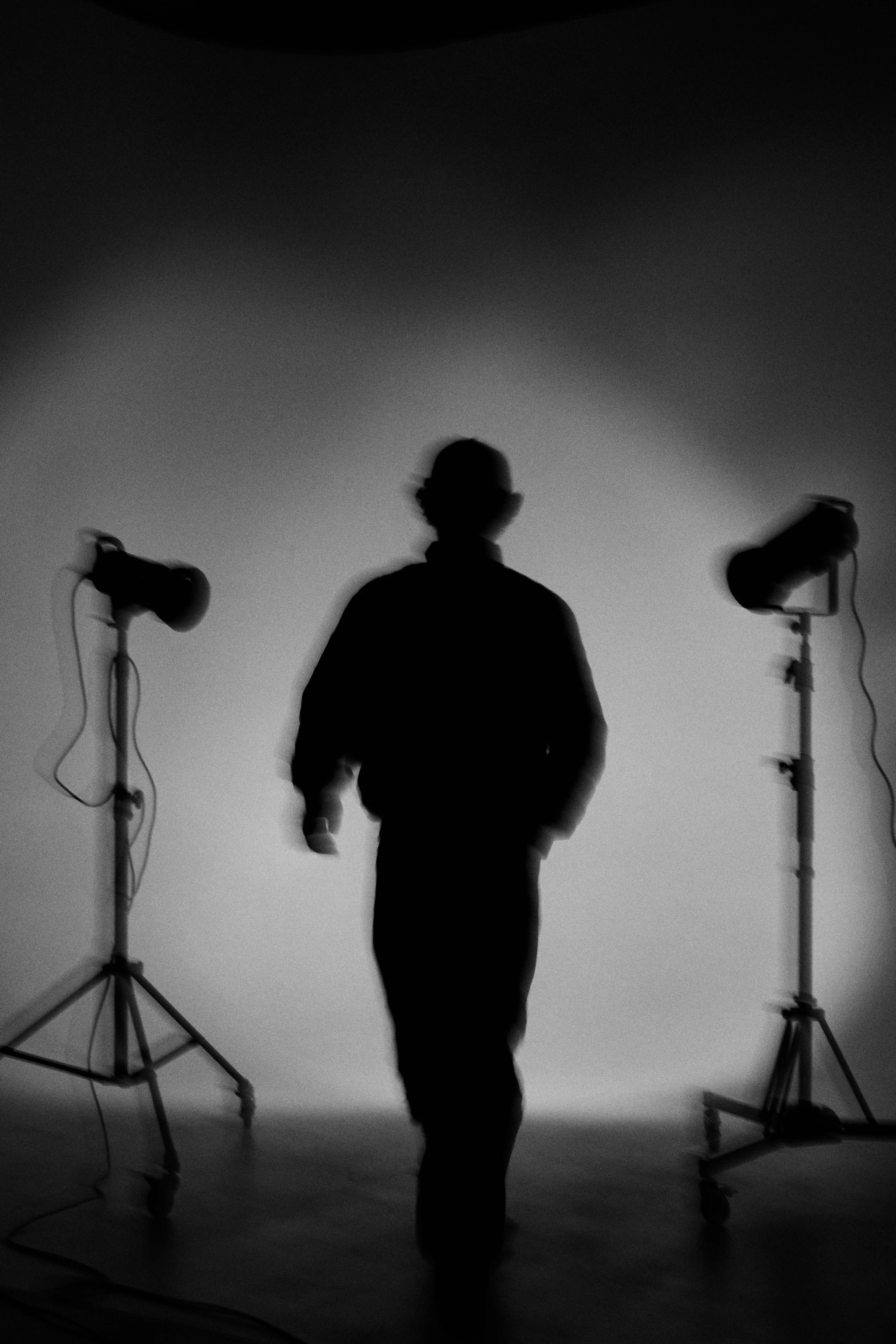 A person in dark clothing standing in front of a plain background, with studio lights on stands to the left and right.