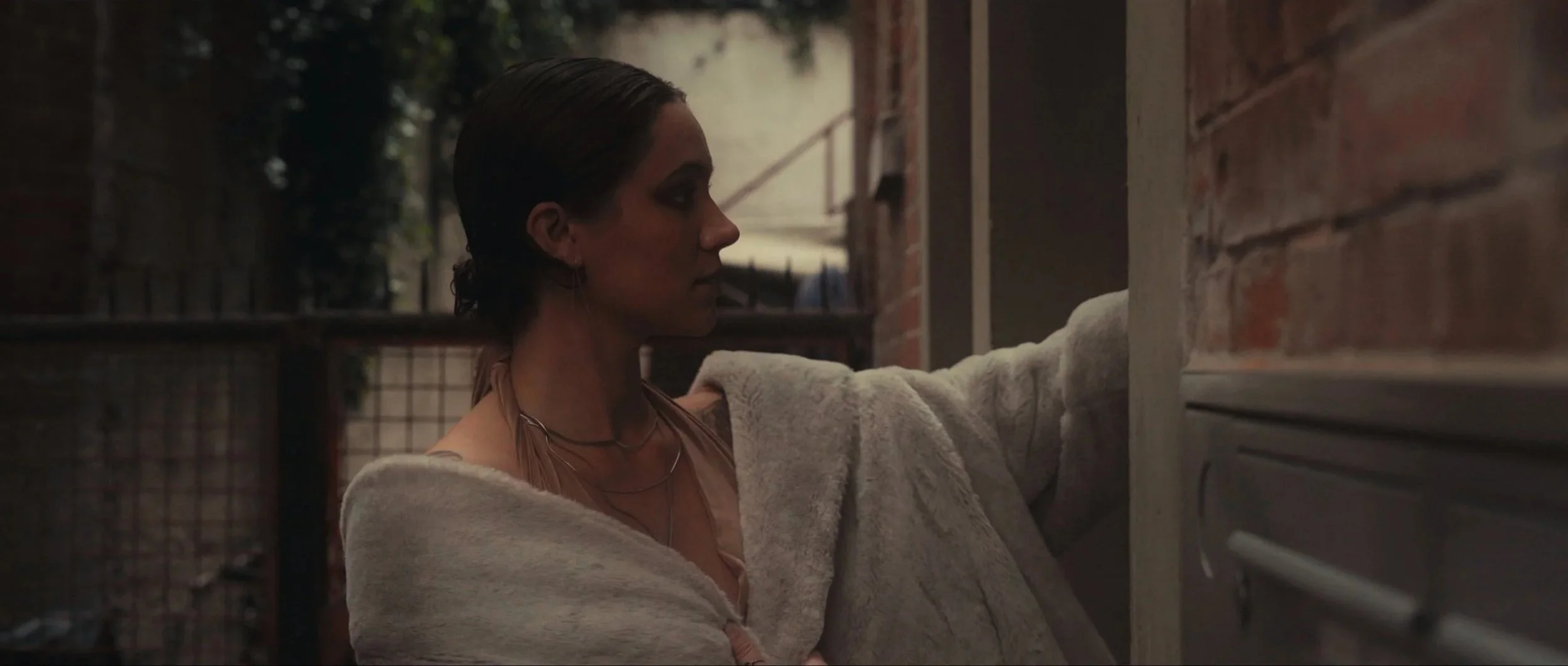 A woman with wet hair, wearing a beige sweater and layered necklaces, standing outdoors near a brick wall, reaching towards the wall with her hand.