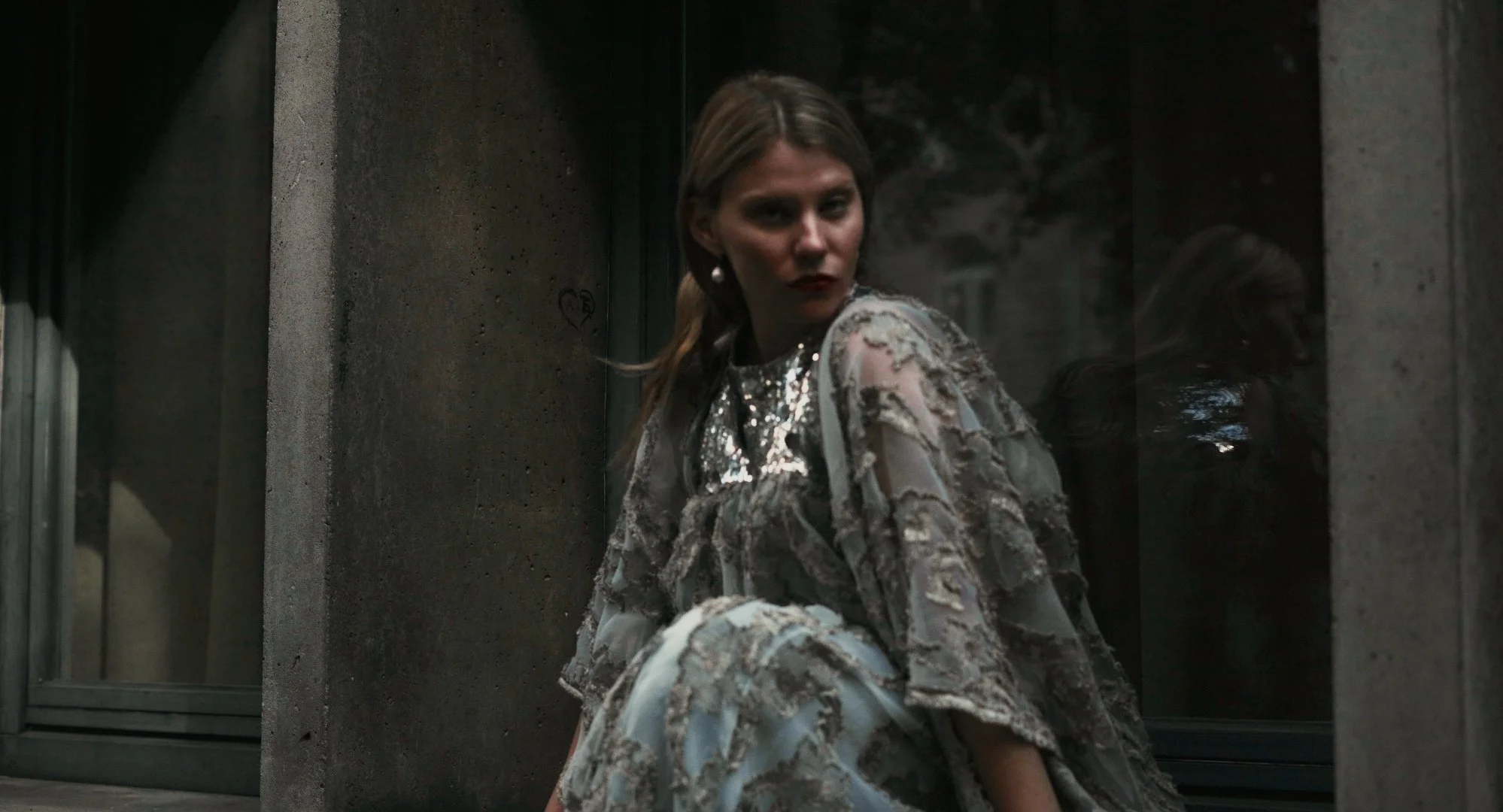 A woman with long hair styled in a side ponytail, wearing a elaborately decorated, shiny, patterned dress, sitting against a dark, industrial-style concrete wall with a reflective surface behind her.