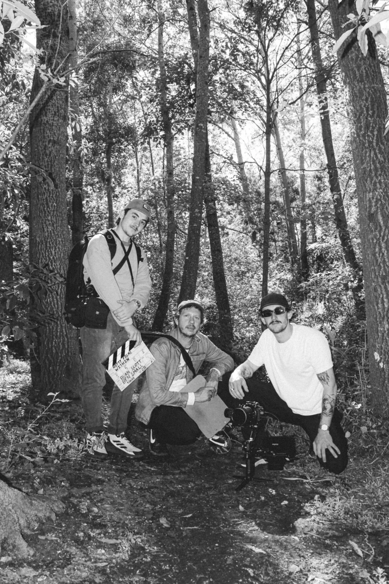 Vier Männer posieren im Wald, ein Mann hält eine Filmklappe, ein weiterer hat eine Kamera. Es sieht aus wie eine Filmszene. Die Männer tragen Freizeitkleidung, Sonnenbrillen und Hüte.