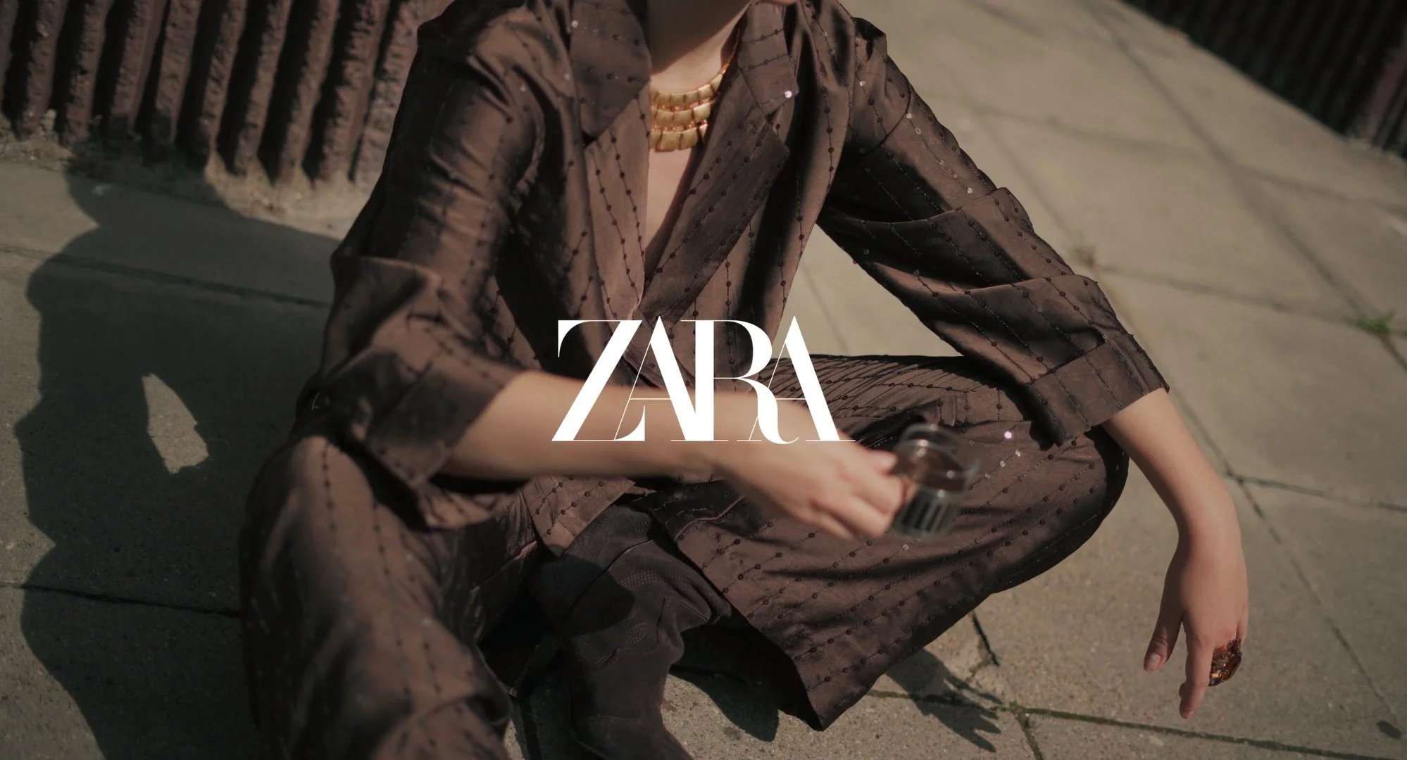 A woman sitting on the ground, wearing a sheer, brown, dotted outfit and a pearl necklace, holding a small object in her hand, with the ZARA logo overlaid.