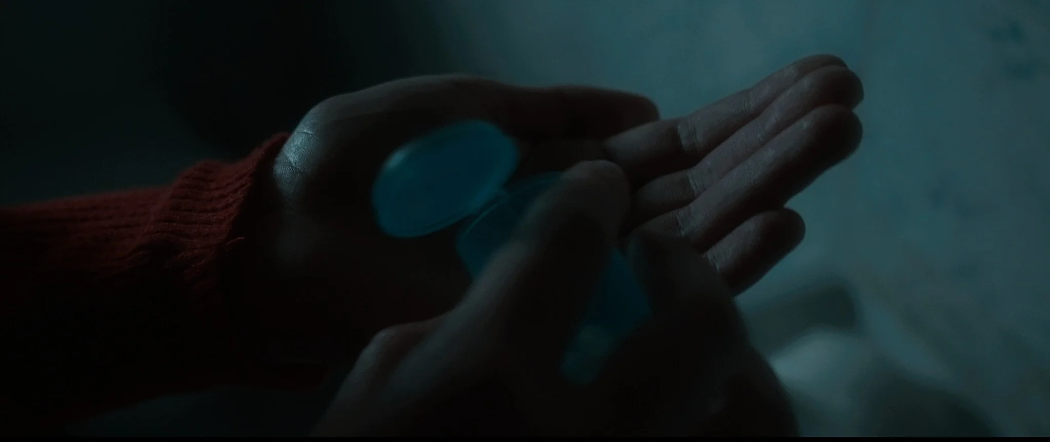 A person's hand holding a small, rounded, opaque blue object in a dimly lit environment.