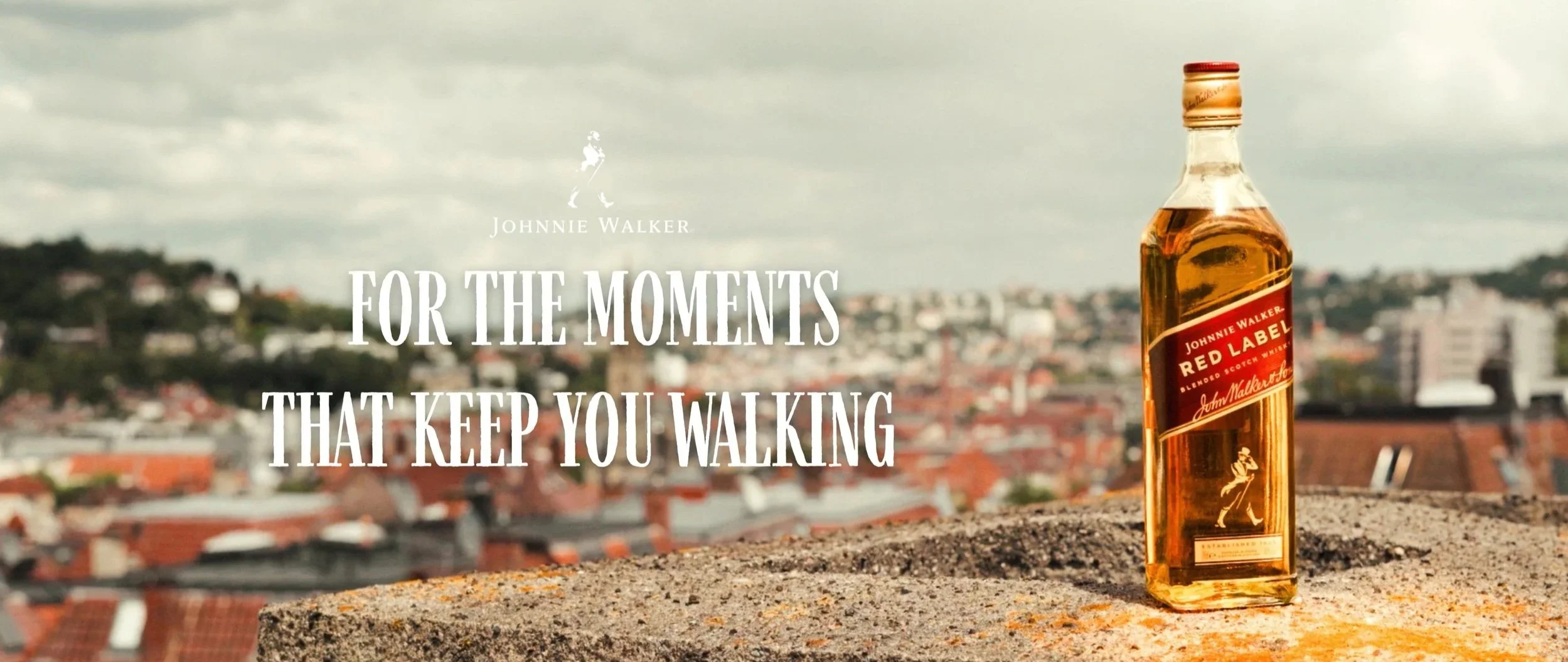 A bottle of Johnnie Walker Red Label blended Scotch whisky on a rooftop ledge with a cityscape in the background and overlaid text reading, 'For the moments that keep you walking.'