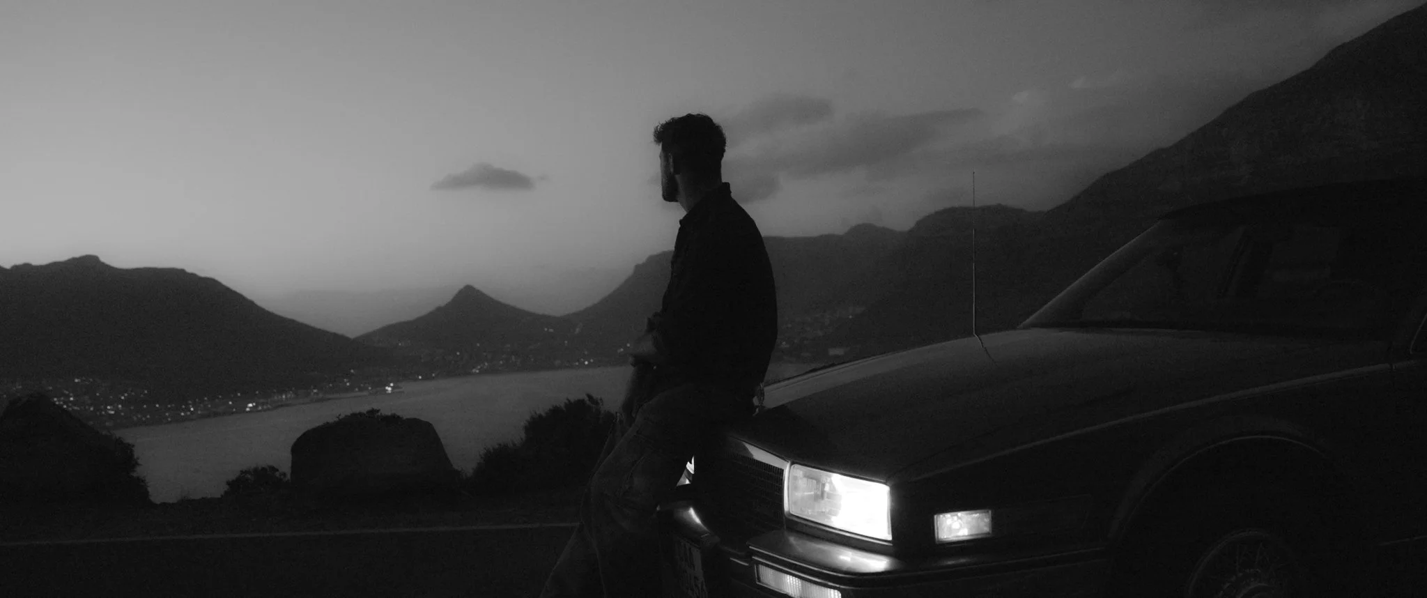 A man stands beside a car on a mountain road during twilight, overlooking a valley with hills and distant lights.