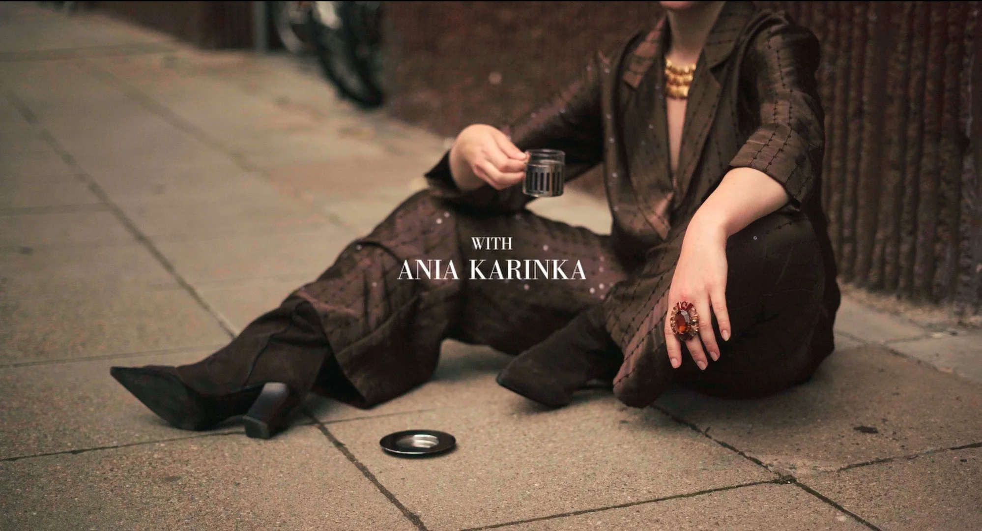 A person dressed in dark, patterned clothing is sitting on the sidewalk against a brick wall, holding a glass with a dark beverage. The person's face is not visible, and they are wearing large statement jewelry. The image has text overlay that says "WITH ANIA KARINKA."