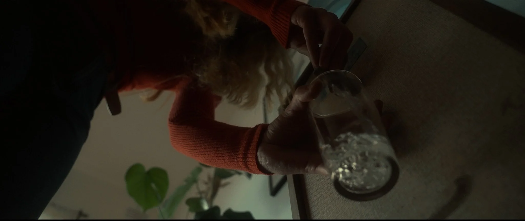 Person with curly blonde hair in a red sweater is reaching for a glass of water on a brown table, with plants visible in the background.