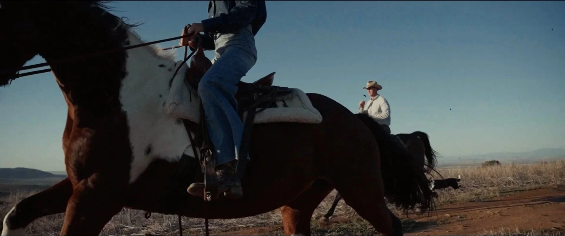 A cowboy riding a horse across an open field with another cowboy standing and eating an ice cream cone in the background.