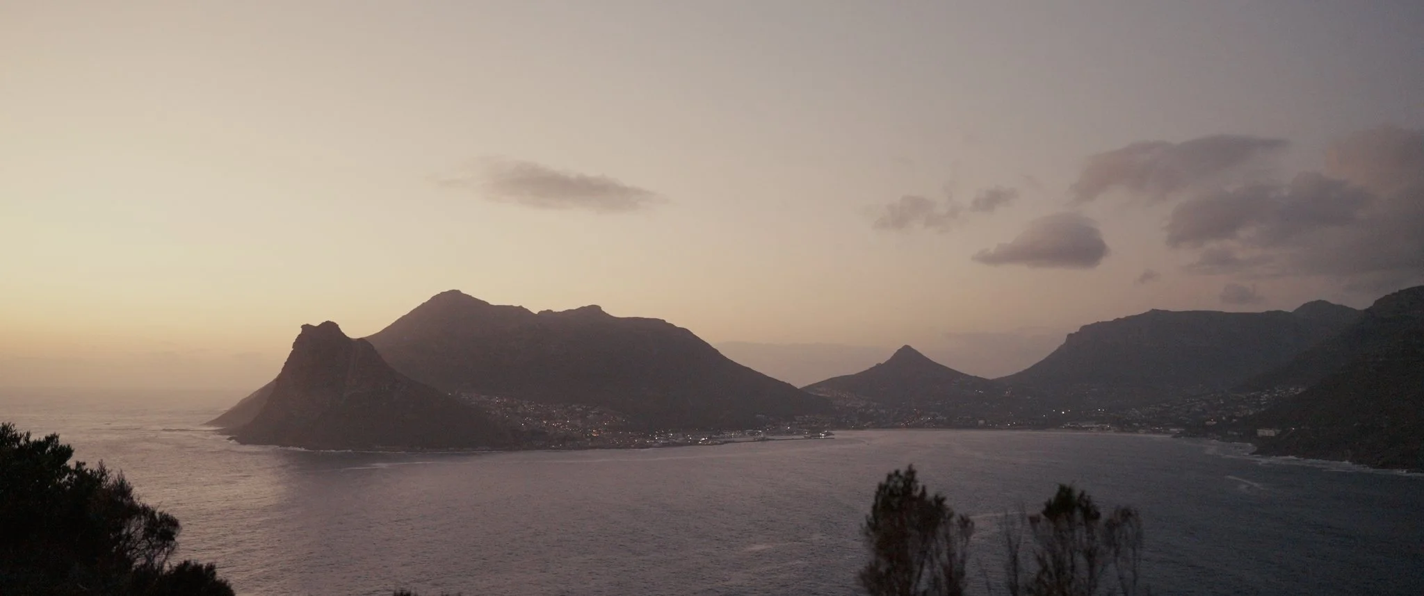 Sunset over a coastal mountain landscape with a calm bay, silhouettes of trees in the foreground, and a partly cloudy sky.