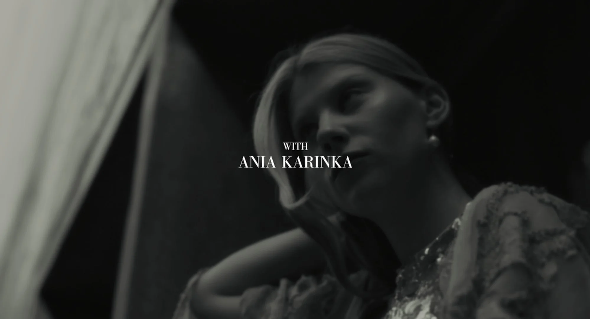 Black and white photo of a woman with glasses and earrings, looking downward, with text overlay that says "WITH ANIA KARINKA".