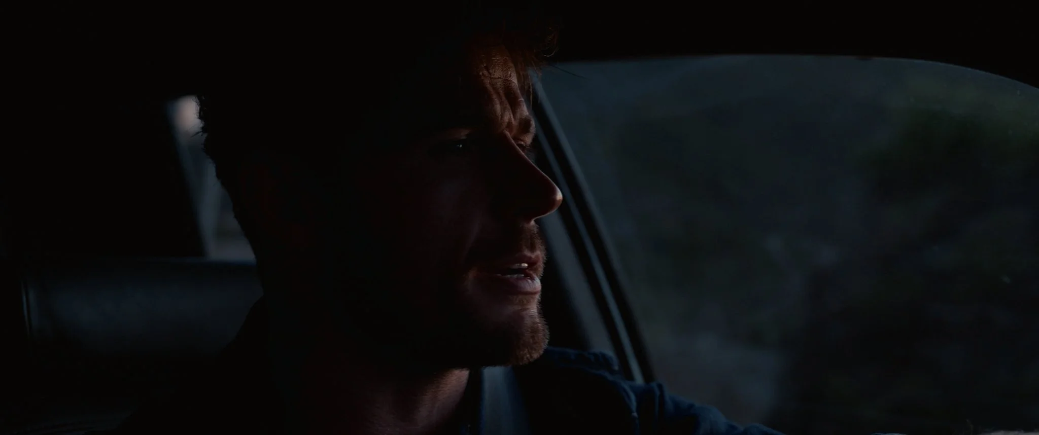 A man with light brown hair and a beard driving a car during dusk or dawn, with his face partially illuminated and a serious expression.