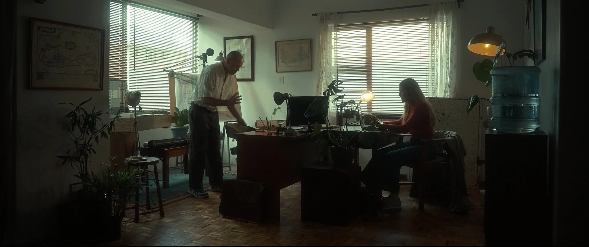 An office with two people, one standing and cleaning a desk, the other sitting and working on a laptop, illuminated by natural light coming through large windows with blinds, surrounded by plants and framed maps on the wall.
