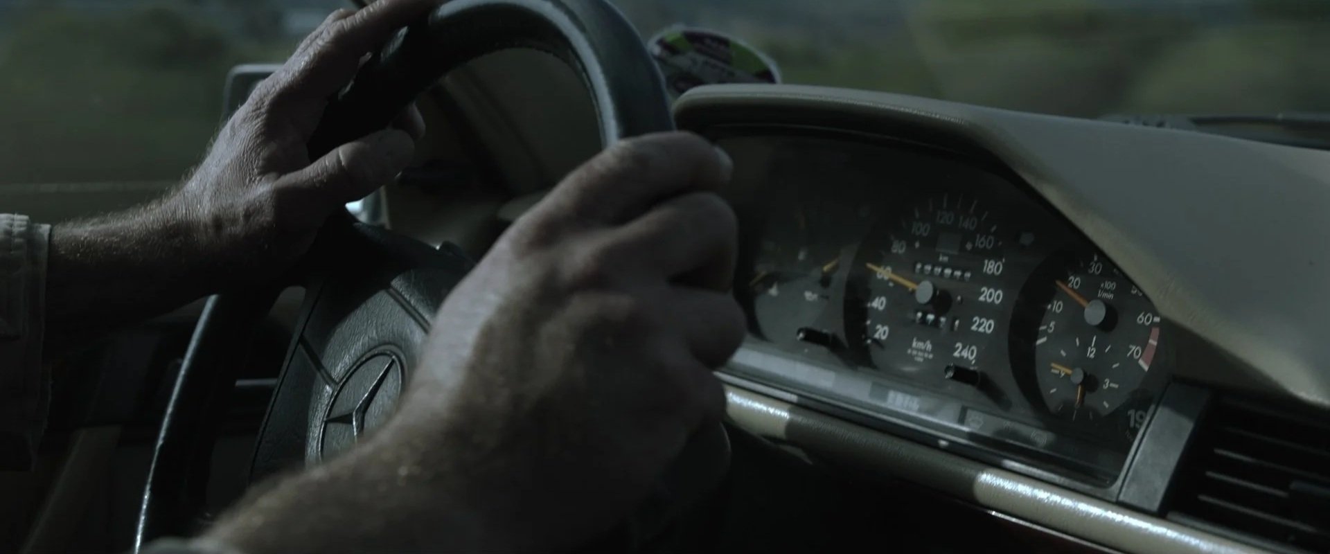 Close-up of person’s hands gripping a steering wheel inside a car, with the dashboard and speedometer visible.