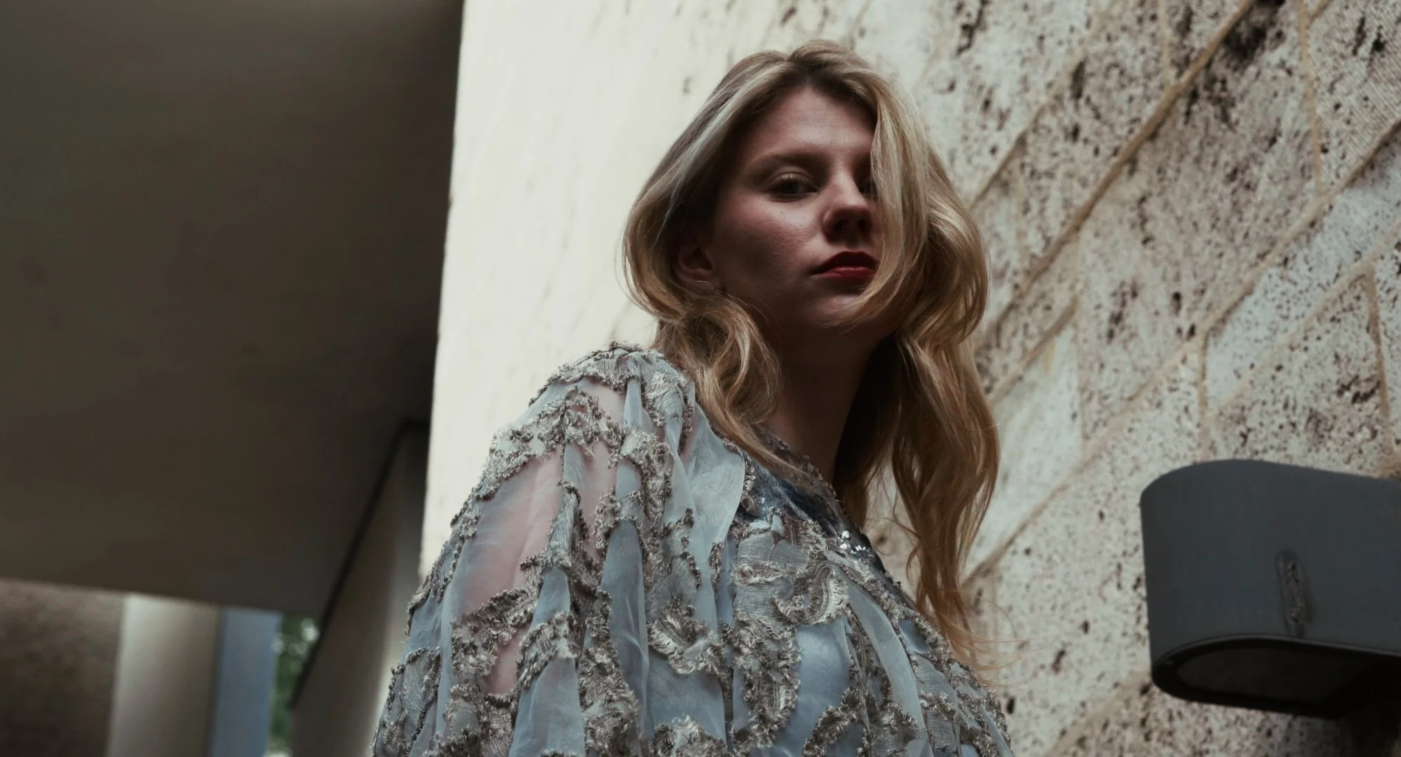 A woman with long, wavy blonde hair leans against a textured brick wall, wearing a light-colored, embroidered dress, looking at the camera.