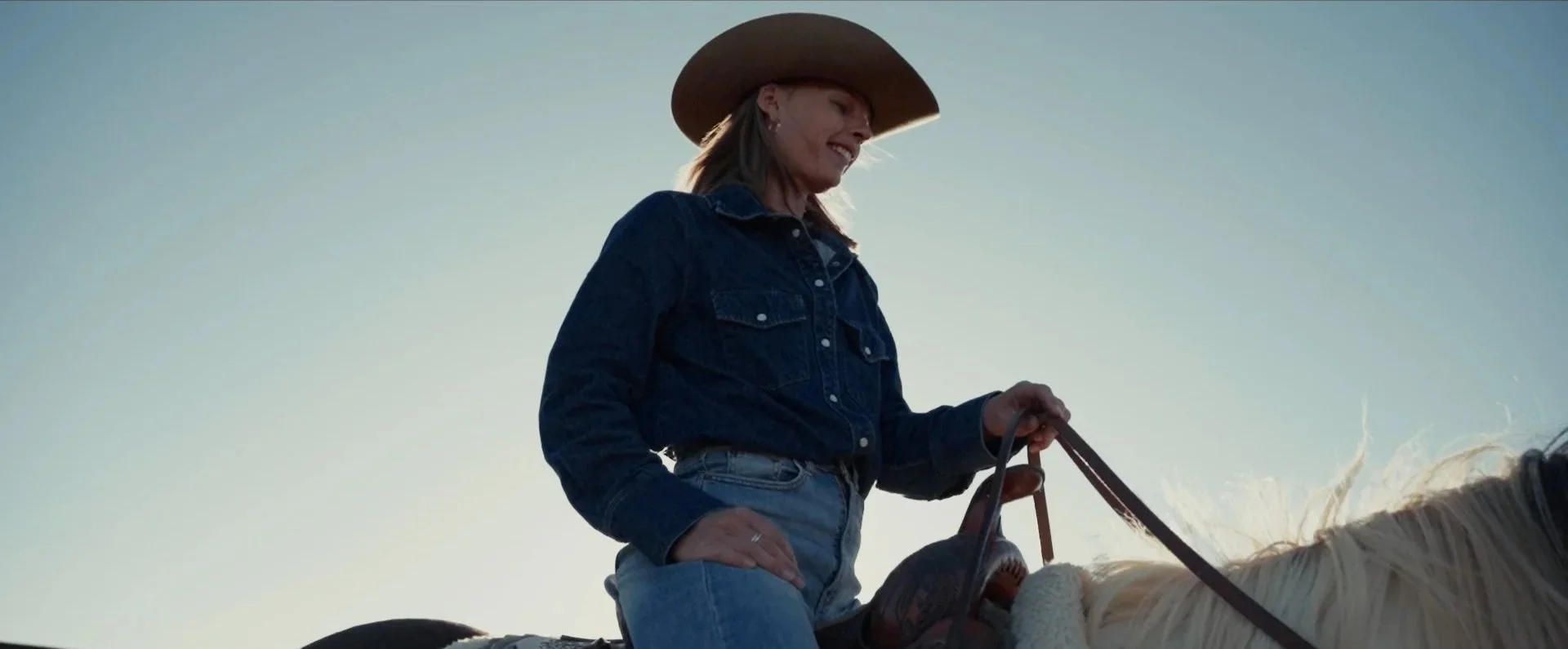 A woman wearing a cowboy hat, denim jacket, and blue jeans riding a horse during daytime.