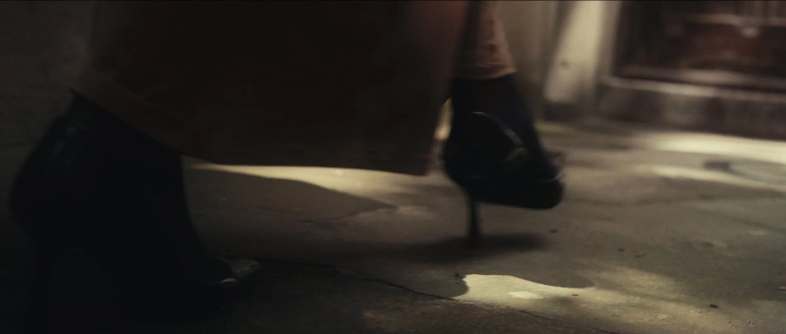 Close-up of a person's feet wearing black high-heeled shoes walking on a dimly lit floor in a possibly public or commercial space.