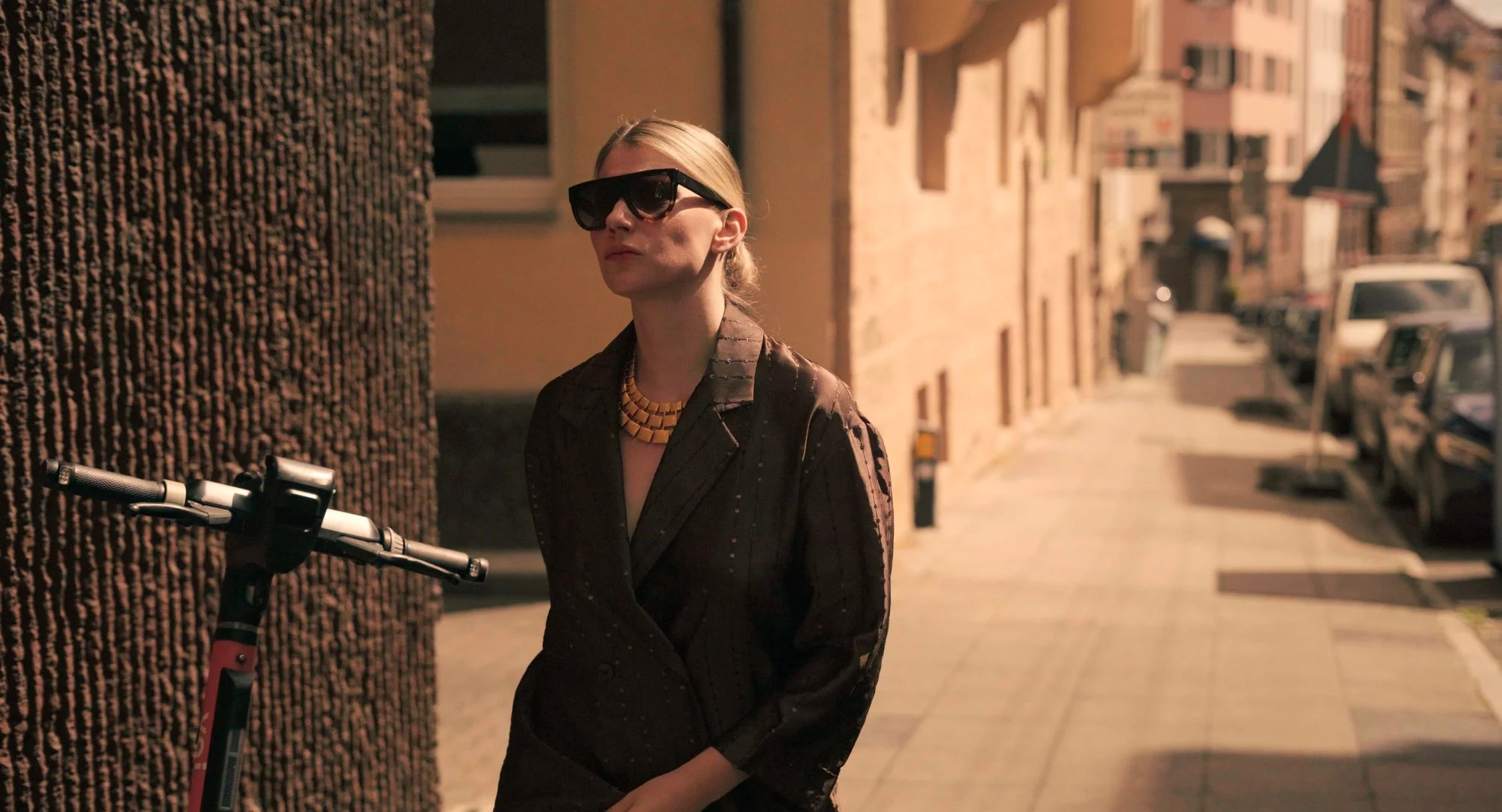 A woman with blonde hair, wearing dark sunglasses and a black coat, stands on a sidewalk next to a parked scooter, in an urban setting during late afternoon or early evening.