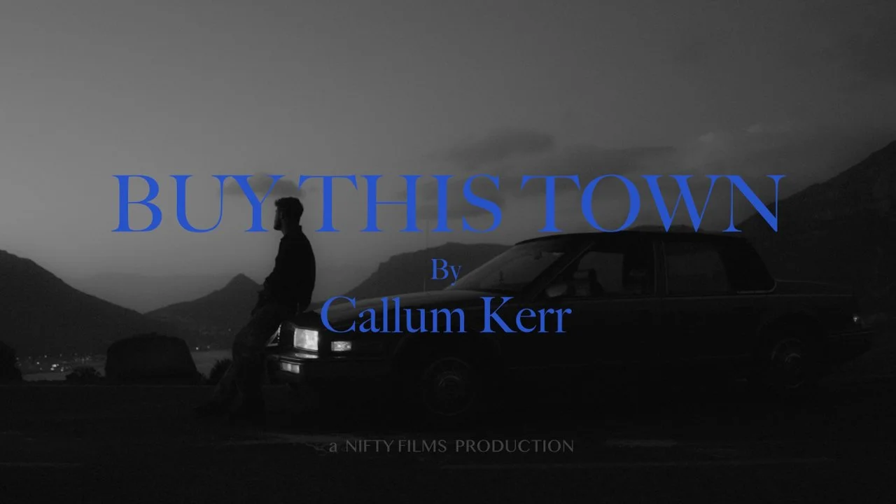 A black and white scene with a man leaning against a car parked on a mountain road. Mountains are visible in the background. Text over the image reads: 'BUY THIS TOWN By Callum Kerr' and at the bottom, 'a NIFTY FILMS PRODUCTION'.