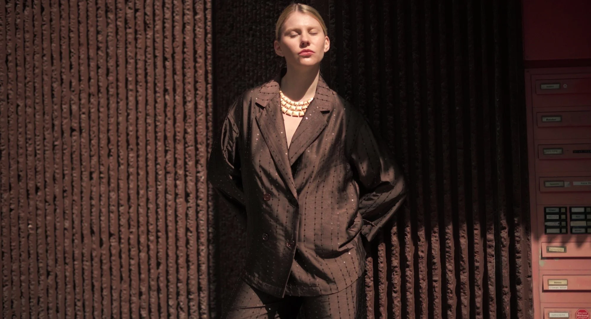 A woman with blonde hair standing with her eyes closed, wearing a brown pinstripe blazer and matching trousers, accessorized with a chunky gold necklace. She is standing in front of a dark textured wall next to pink mailboxes.