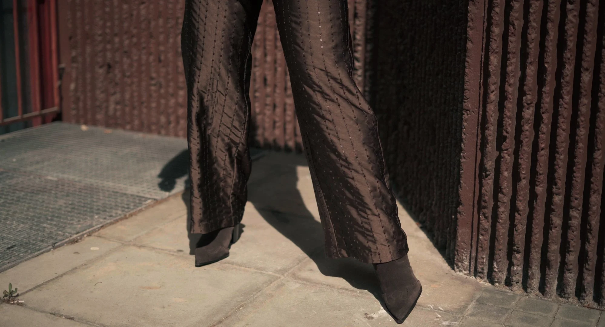 Close-up of a person's legs wearing sheer, black, sparkly pants and black heeled boots, standing on a sidewalk next to a textured dark brown wall.