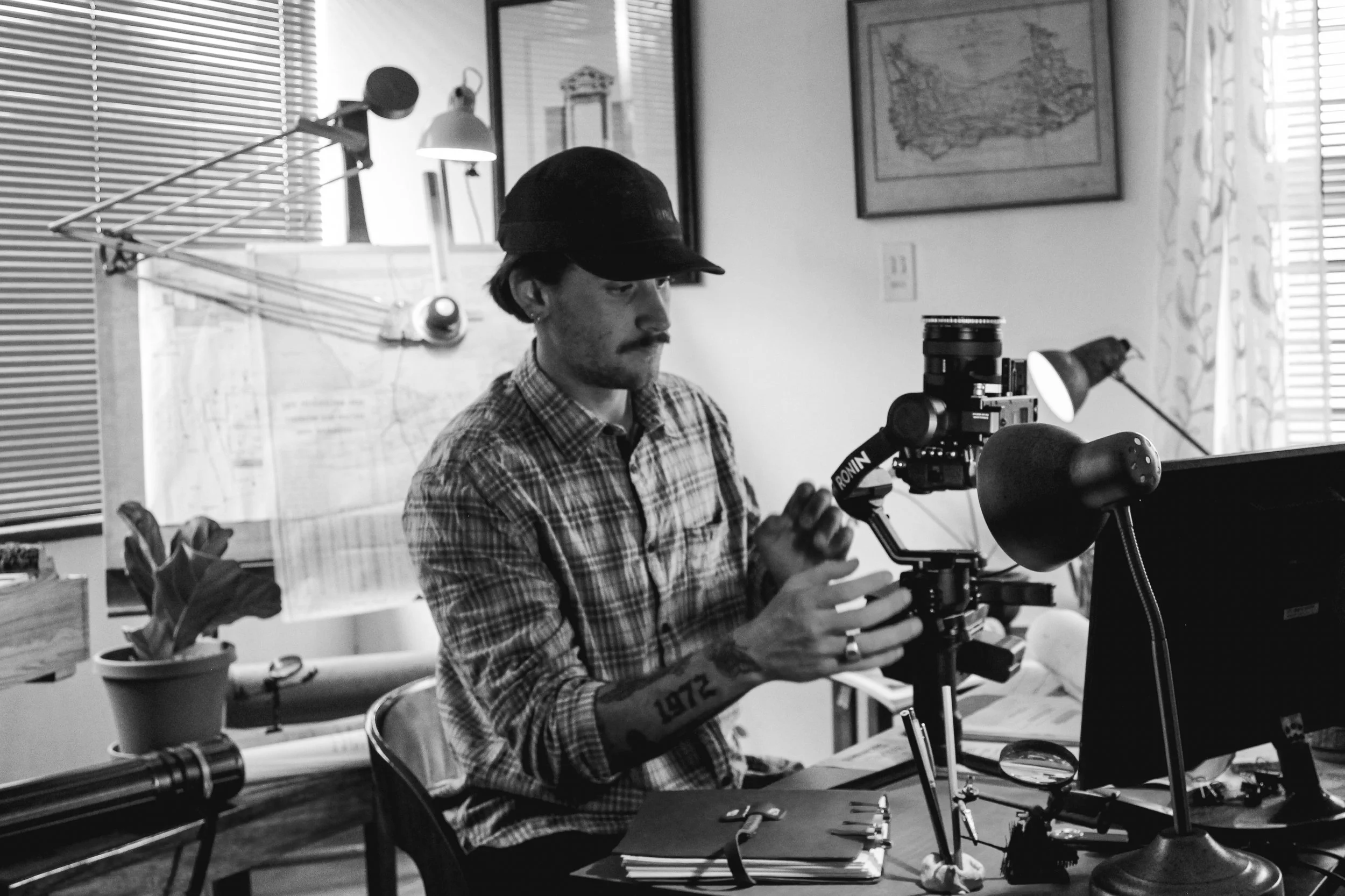 Ein Mann mit dunklem Hut und kariertem Hemd arbeitet an einem Mikroskop in einem Büro, im Hintergrund befinden sich Fenster mit Vorhängen, Wandbilder und Schreibwaren auf einem Tisch.