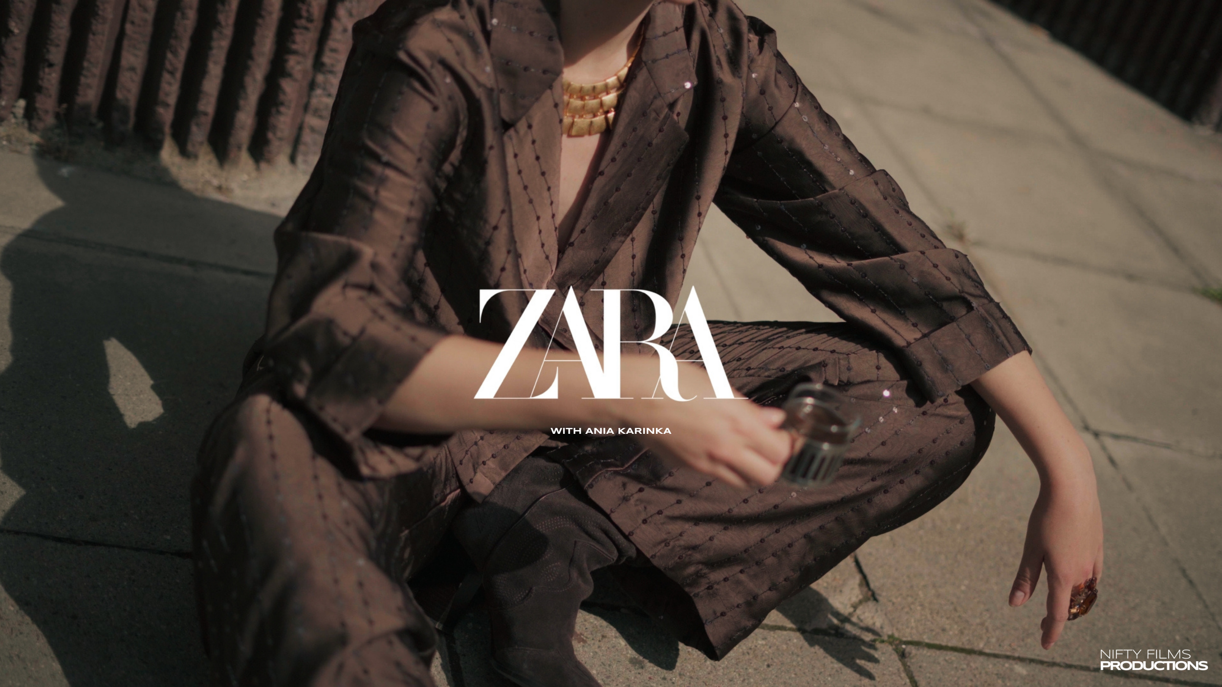 Person sitting on the sidewalk, wearing a brown outfit with a necklace, holding a bottle, with a fence in the background.