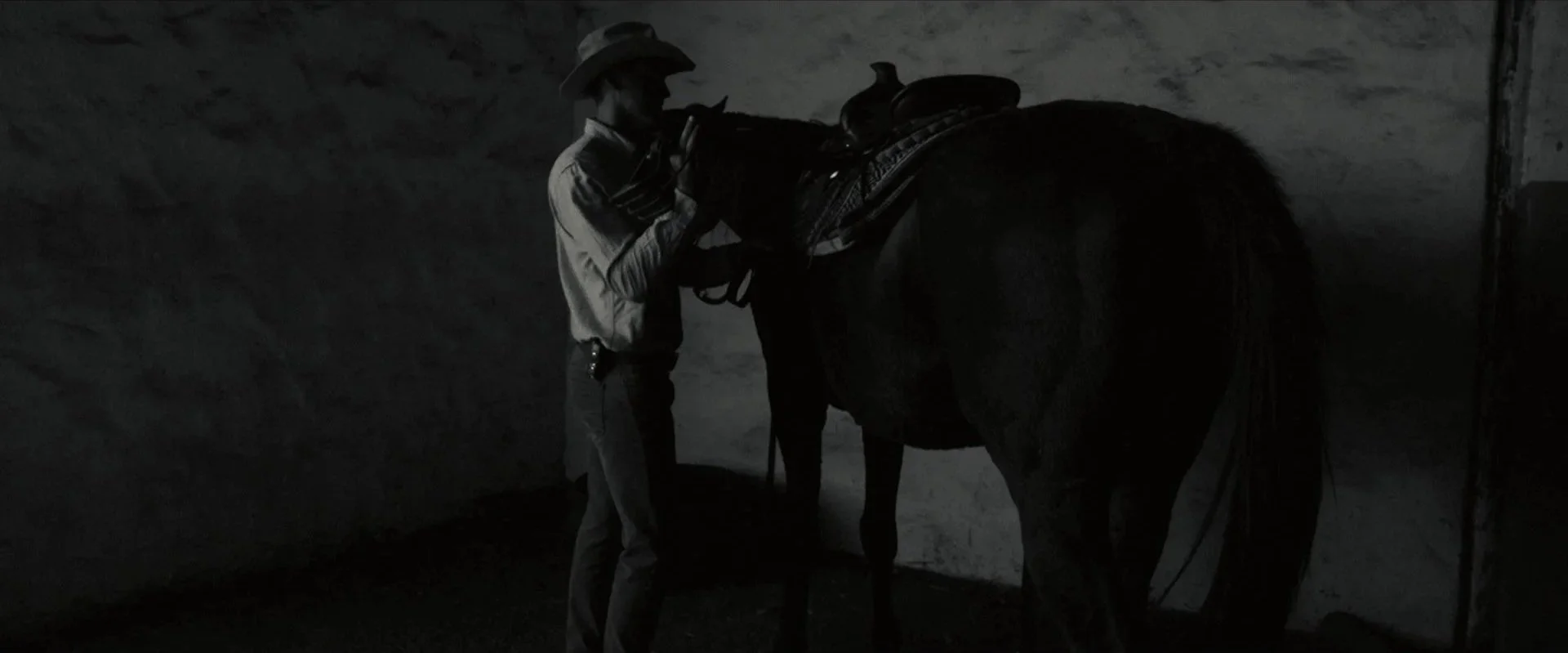 Dark photo of a man in western attire grooming a horse in a dimly lit environment.