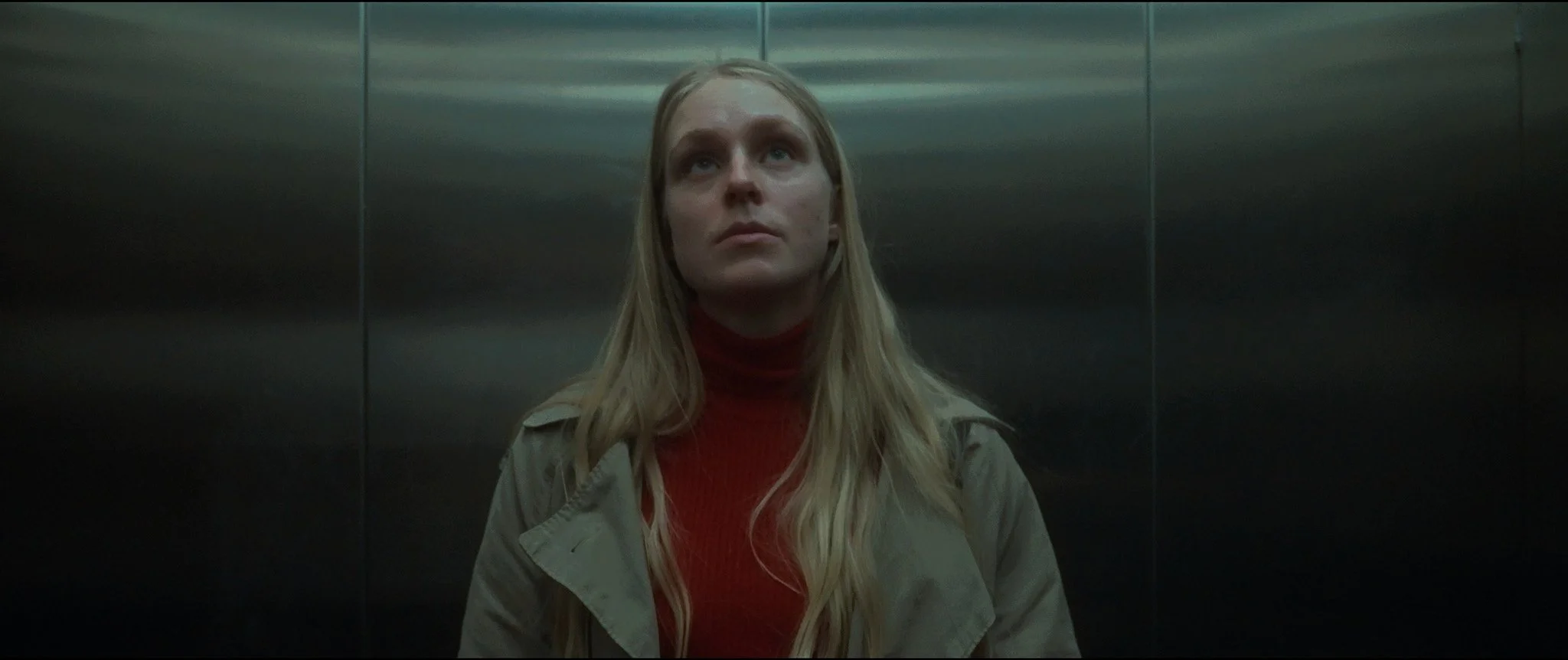A woman standing in an elevator, looking upward with a neutral expression, wearing a beige jacket and a red turtleneck, with stainless steel interior walls.