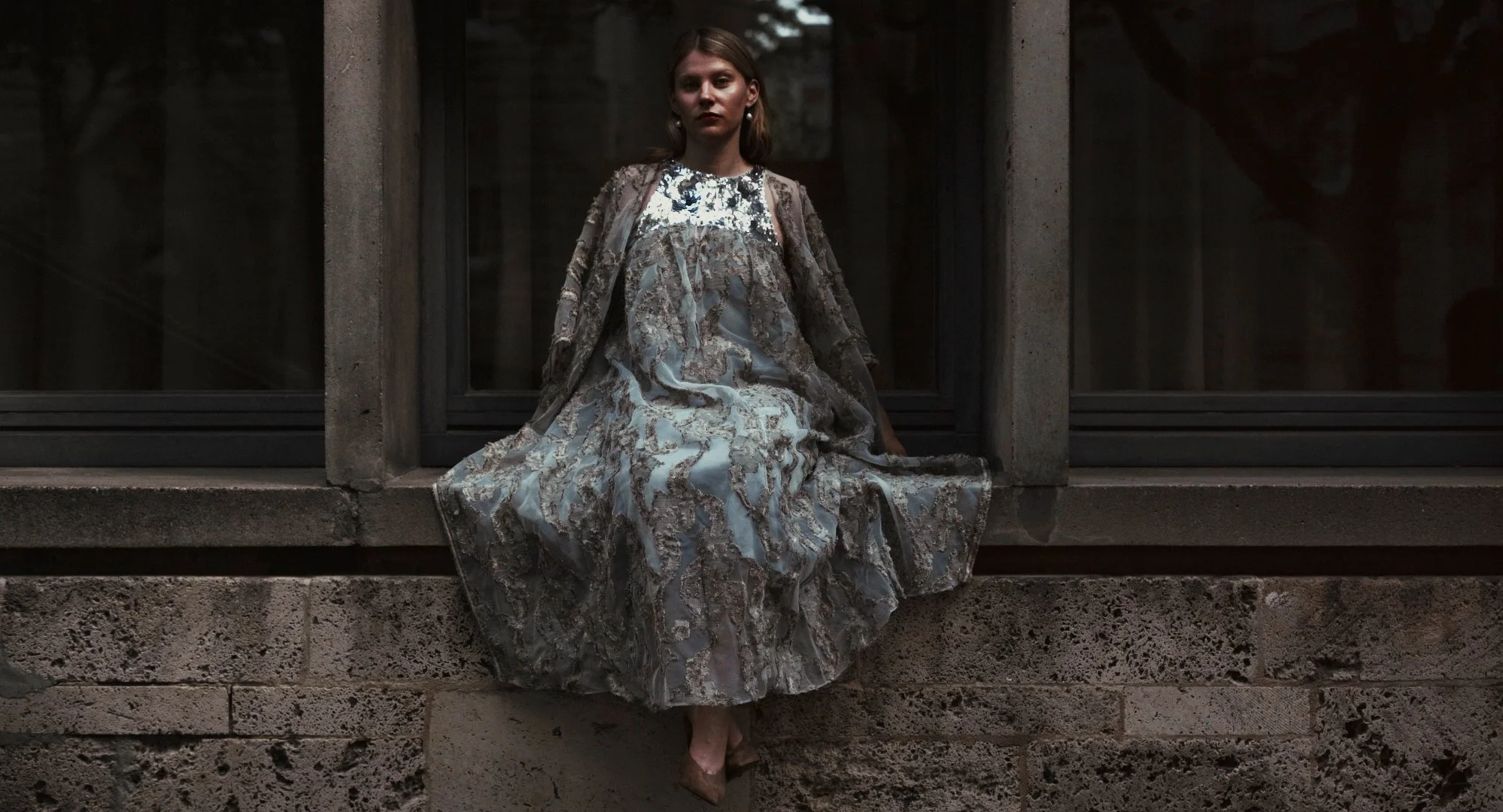 woman in a silver, patterned dress sitting on a concrete ledge outside a building with large windows