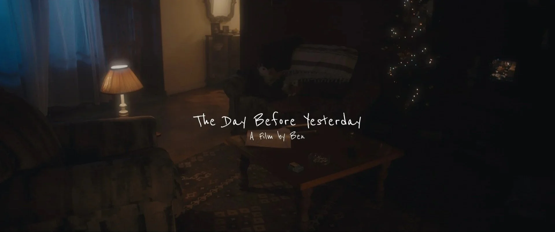 Dimly lit living room with a sofa, wooden coffee table, and a Christmas tree. The title 'The Day Before Yesterday' and the subtitle 'A film by Ben' are overlaid on the image, indicating the opening scene of a film.