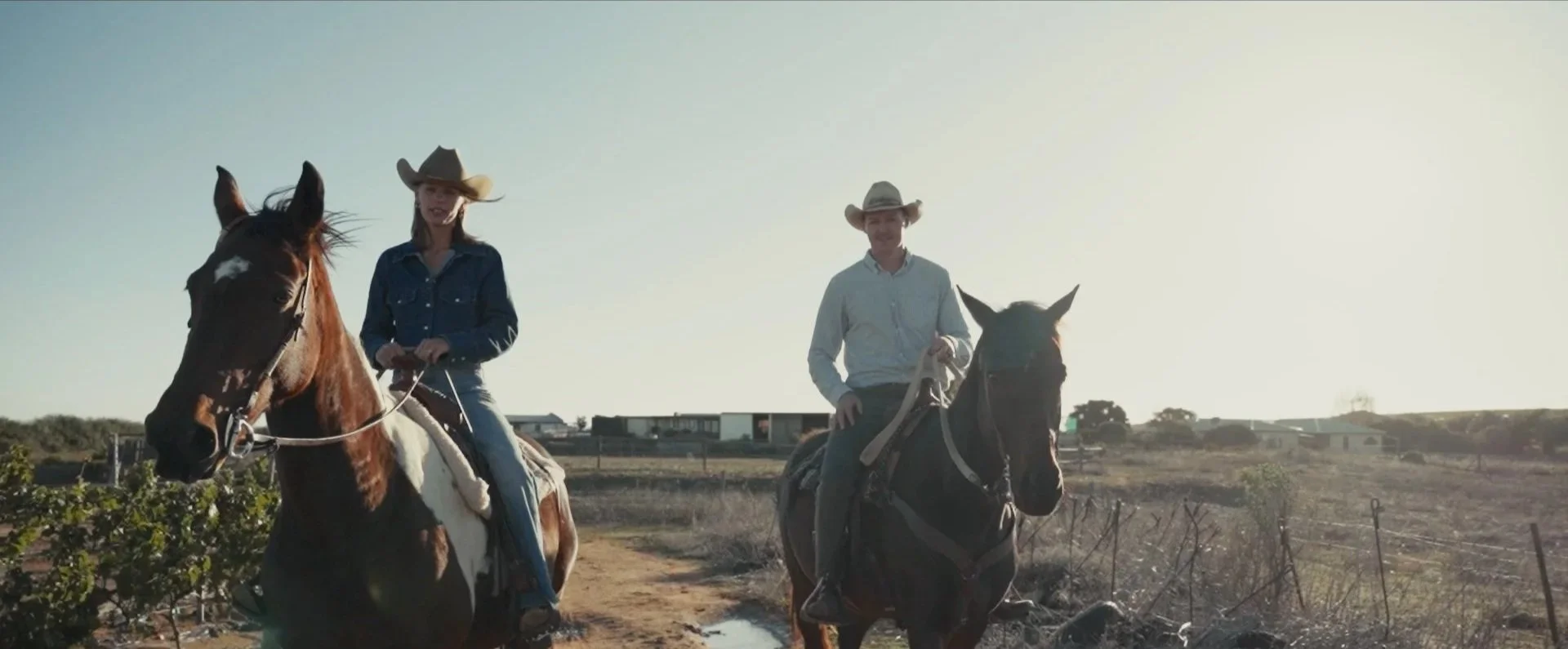 Two people riding horses in an open rural area with clear sky and sparse vegetation.