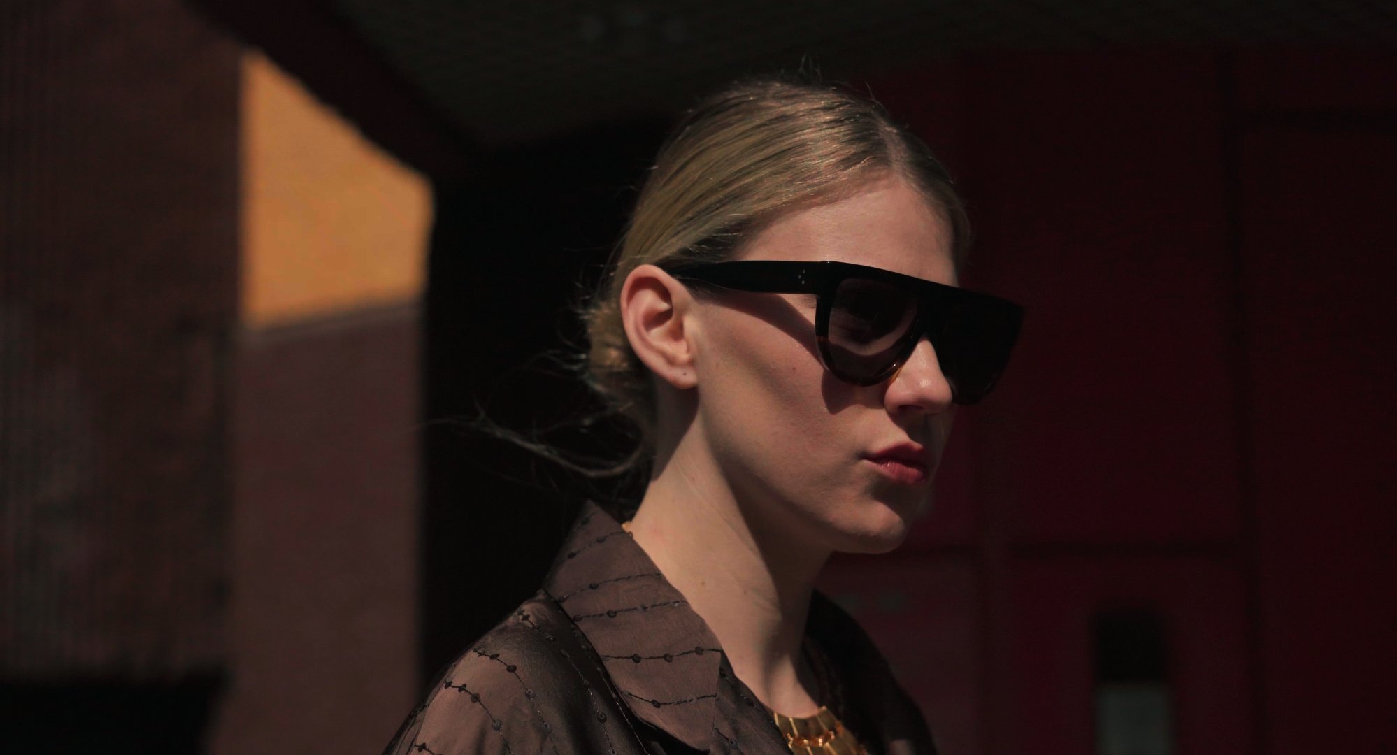 A woman with blonde hair wearing large black sunglasses and a dark, patterned top, standing in a dimly lit environment.