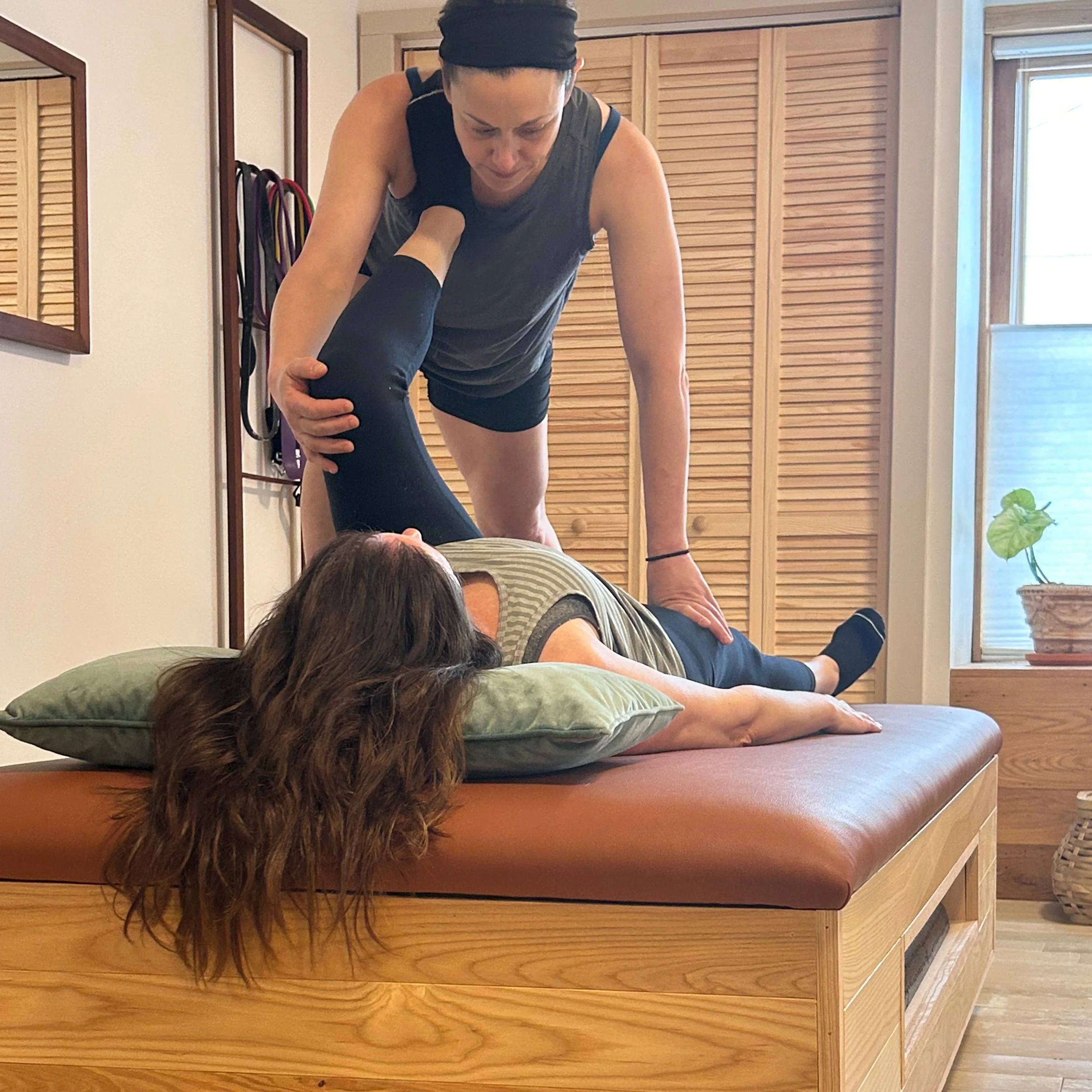 A woman is practicing acro yoga on a wooden bed with her legs and arms extended and her head on the mattress inside a bright room with wooden flooring, a wooden door, and some plants.