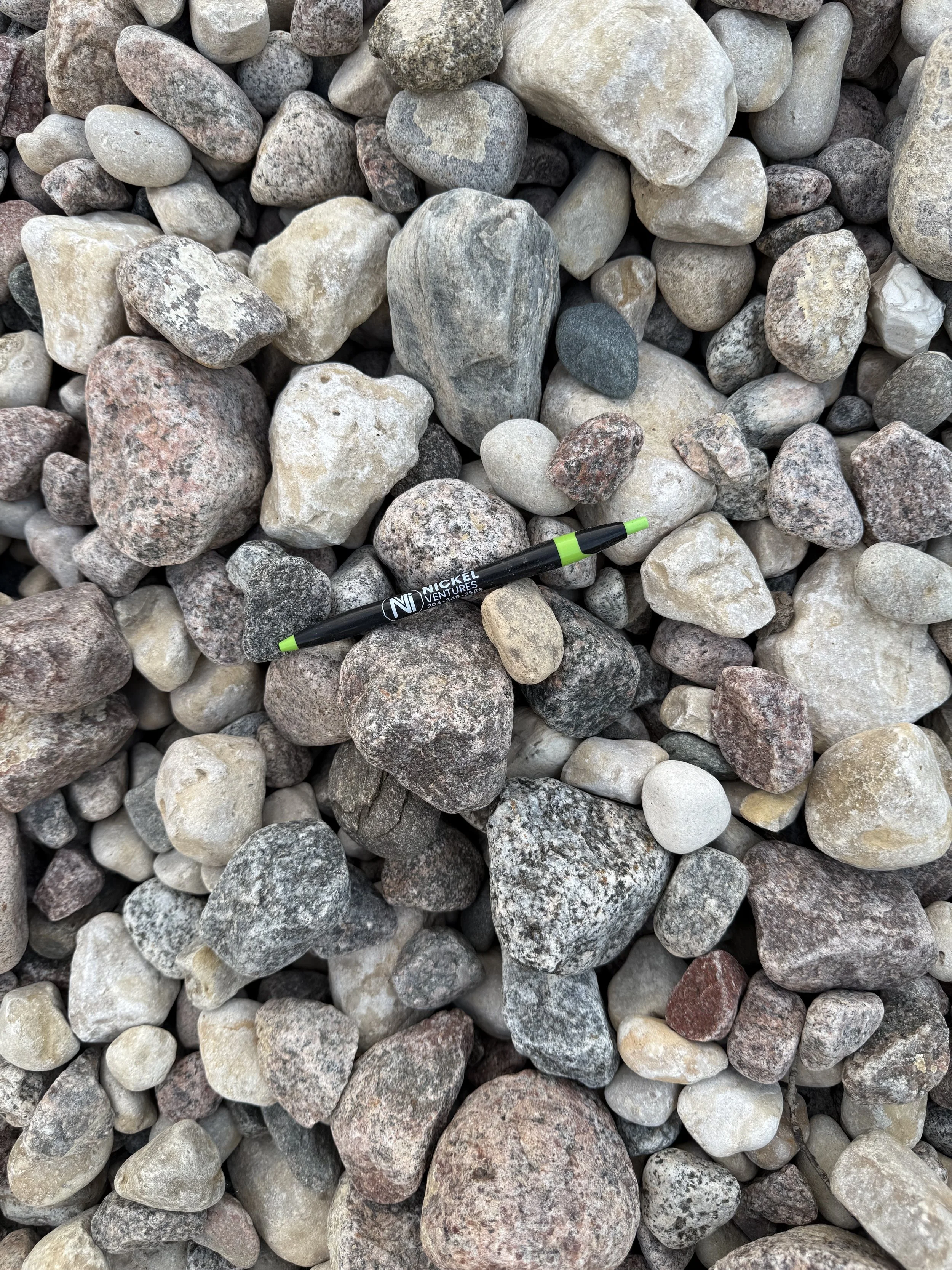 A collection of various small to medium-sized rocks of different colors, textures, and shapes, with a black and green pen lying among them.