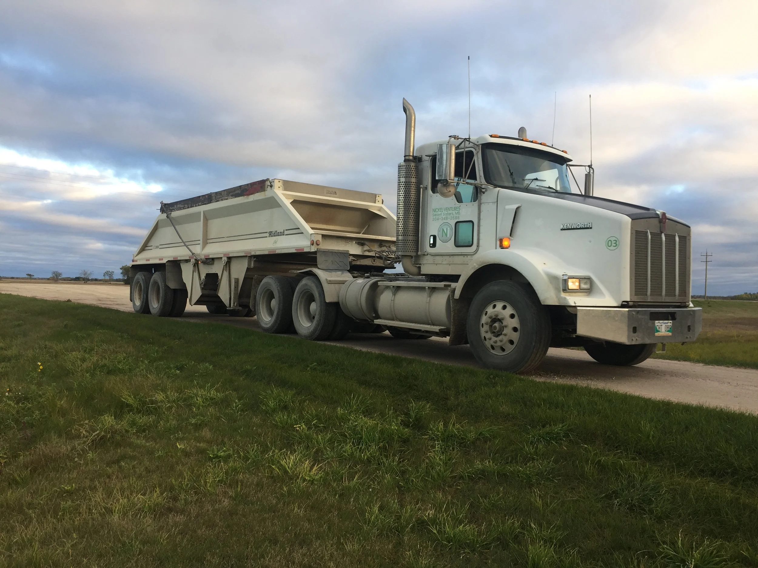 A large white Kenworth truck with a gray hood is parked on a grassy area under a cloudy sky. The truck has multiple antennas and is pulling a belly dump trailer.