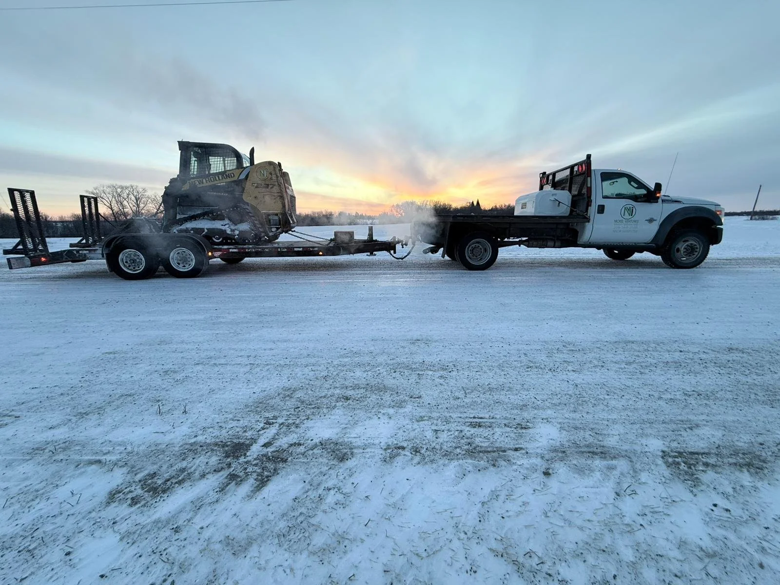 A white pickup truck towing a trailer with a snow groomer attached on a snowy landscape during sunset.