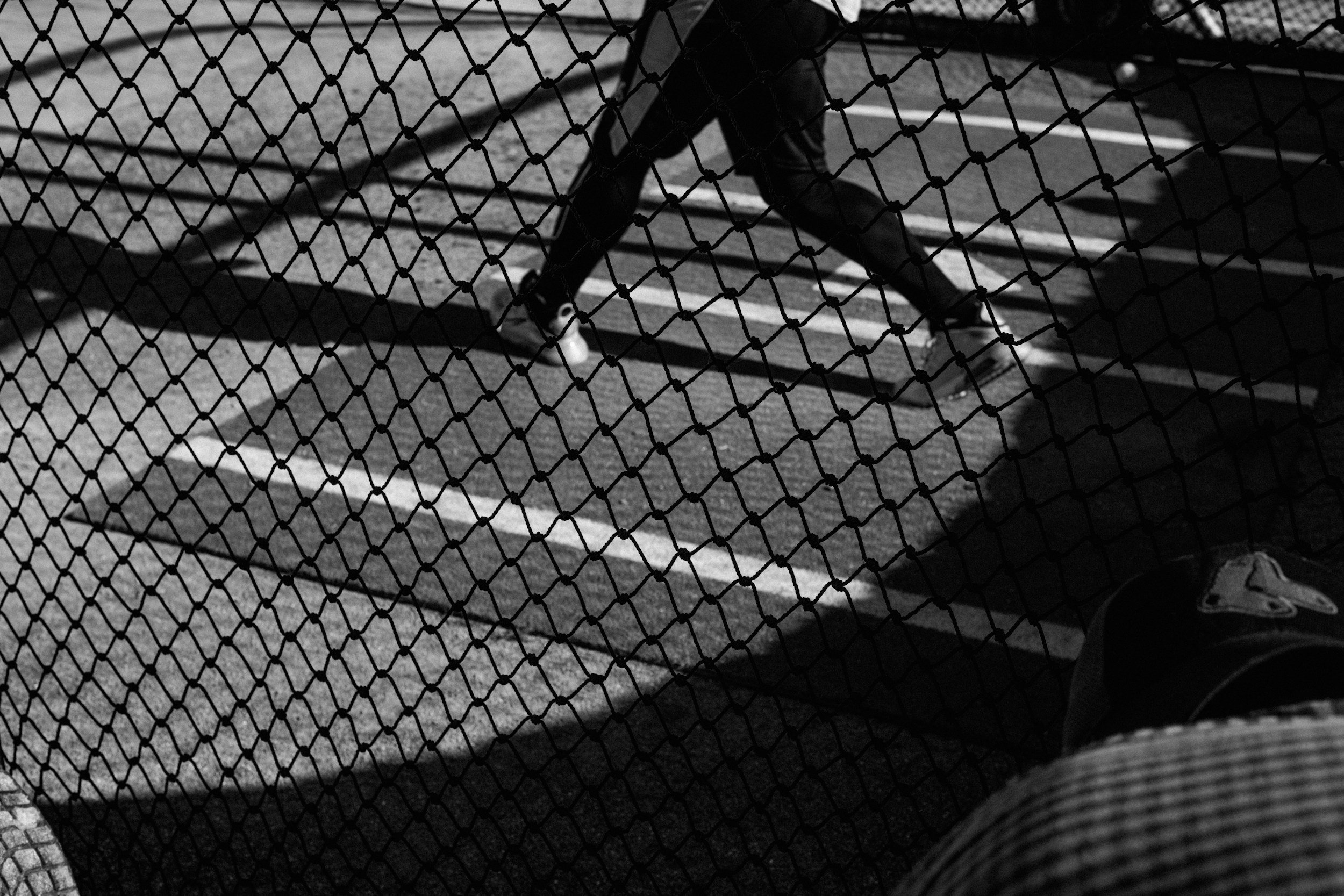 A person skating on a skatepark with a chain-link fence in the foreground, shadows cast on the ground, in black and white.