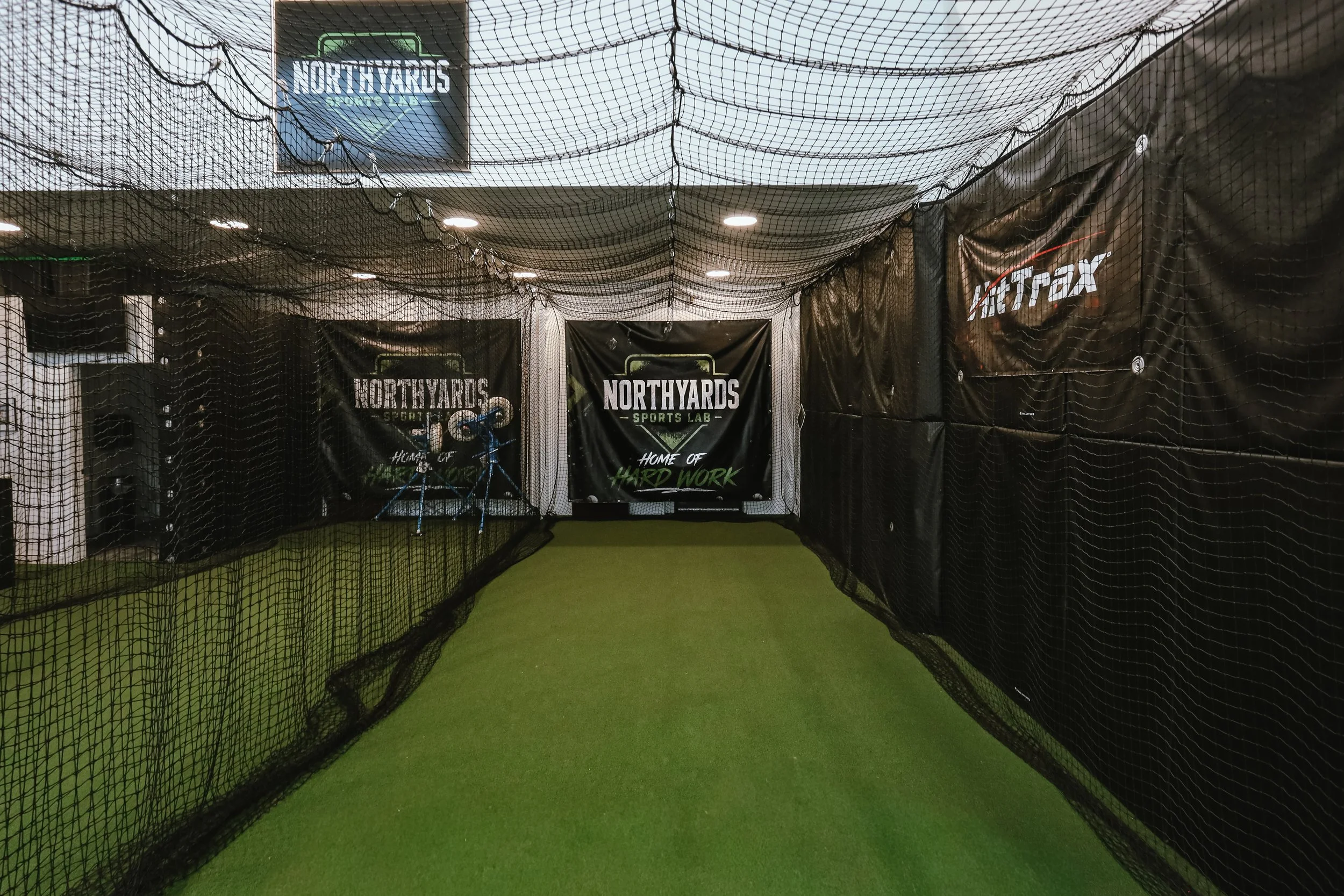 Indoor baseball or softball practice cage with artificial turf, pitching machines, and a monitor displaying a baseball field. Four buckets of softballs and baseballs are on the floor.