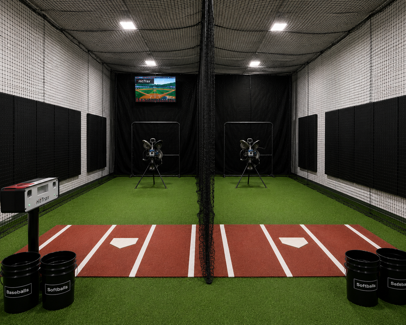 Indoor baseball or softball practice cage with artificial turf, pitching machines, and a monitor displaying a baseball field. Four buckets of softballs and baseballs are on the floor.