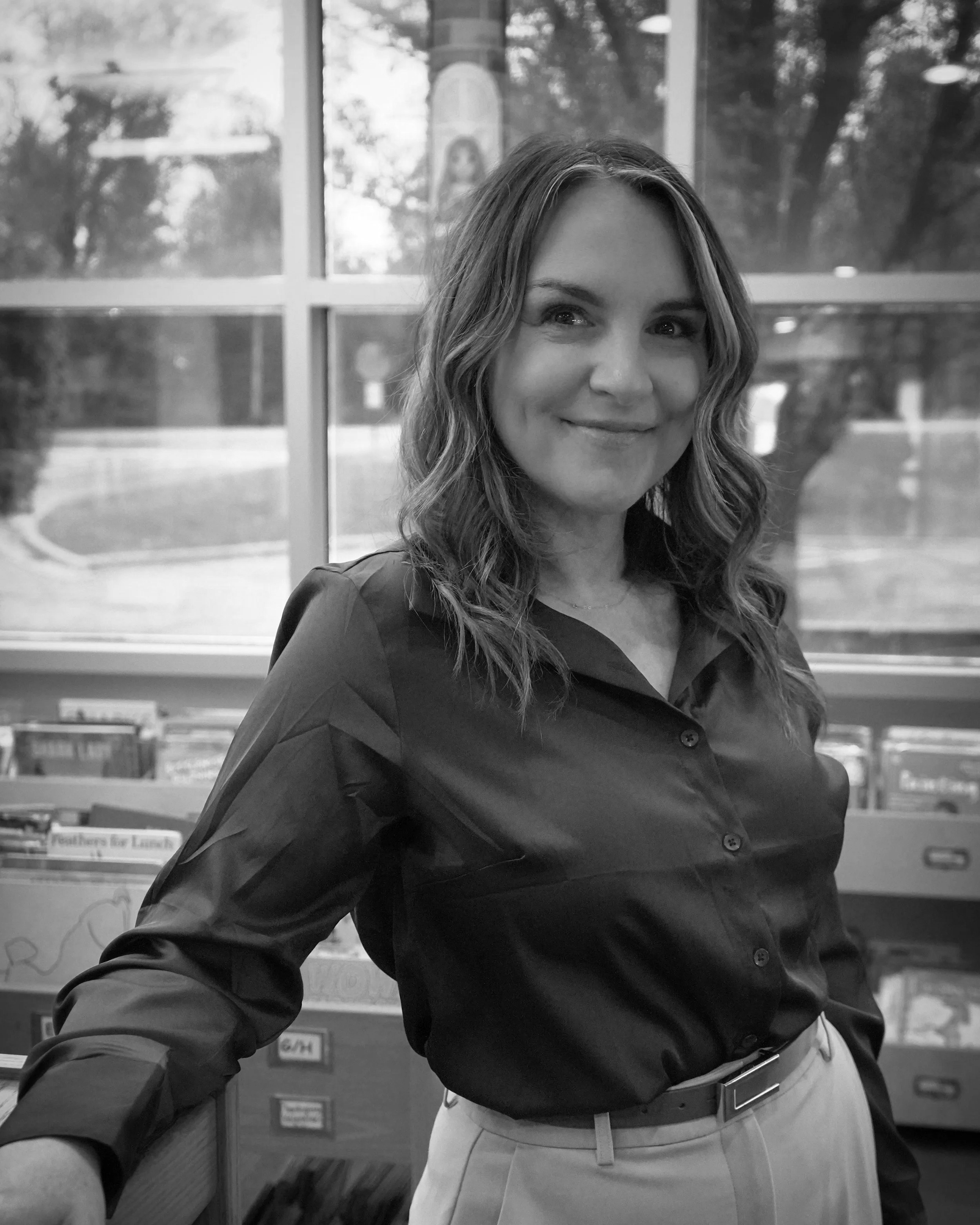 A woman with long, wavy hair smiling and looking slightly to the side, wearing a dark button-up shirt and khaki pants with a belt, standing in front of a large window with blurred greenery and a church visible outside.