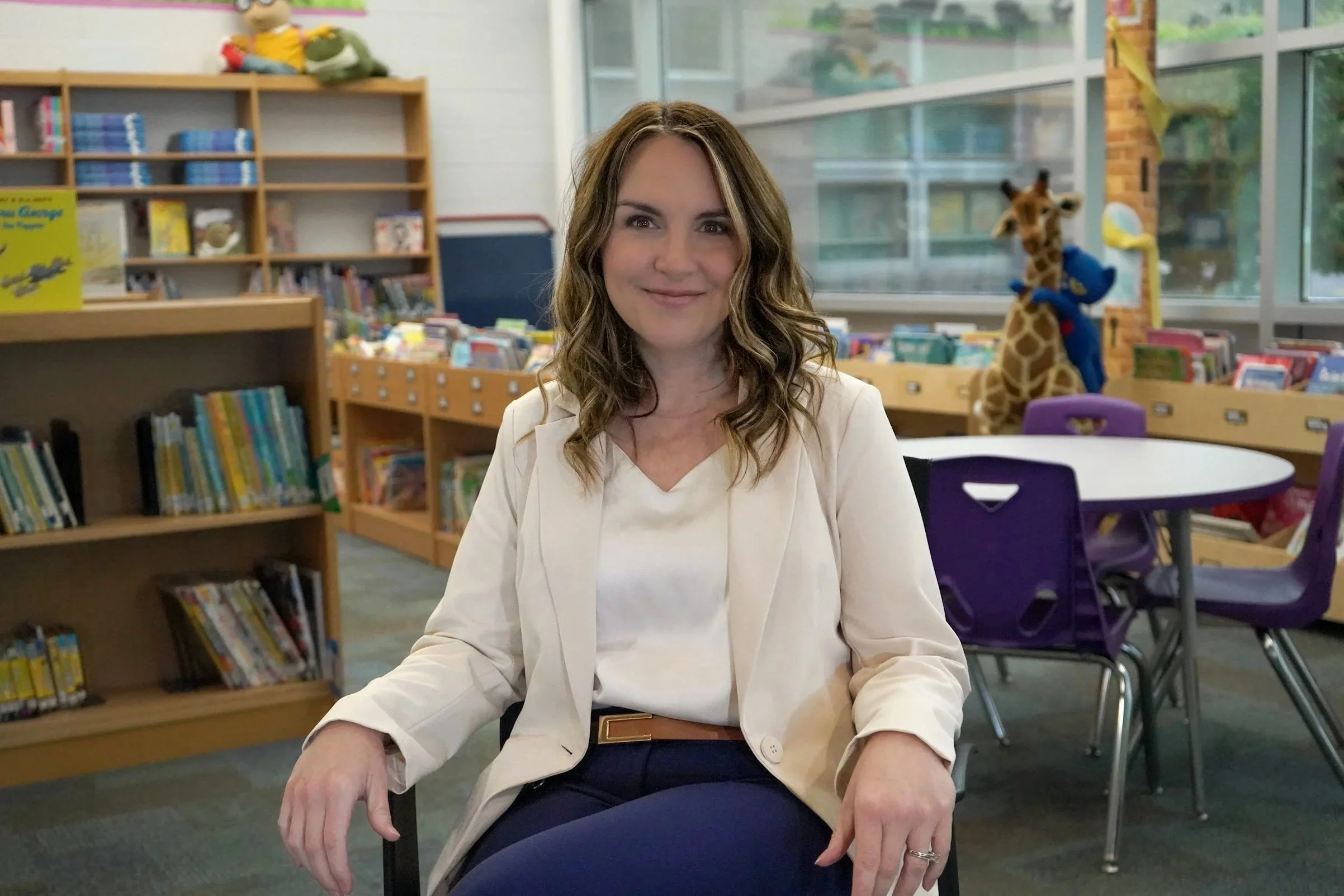 A woman with long wavy brown hair, wearing a white blazer over a white top, sitting at a table in a library or bookstore, smiling slightly at the camera. In the background, there are bookshelves filled with books, a stuffed animal of a giraffe with a blue scarf, and large windows letting in natural light.