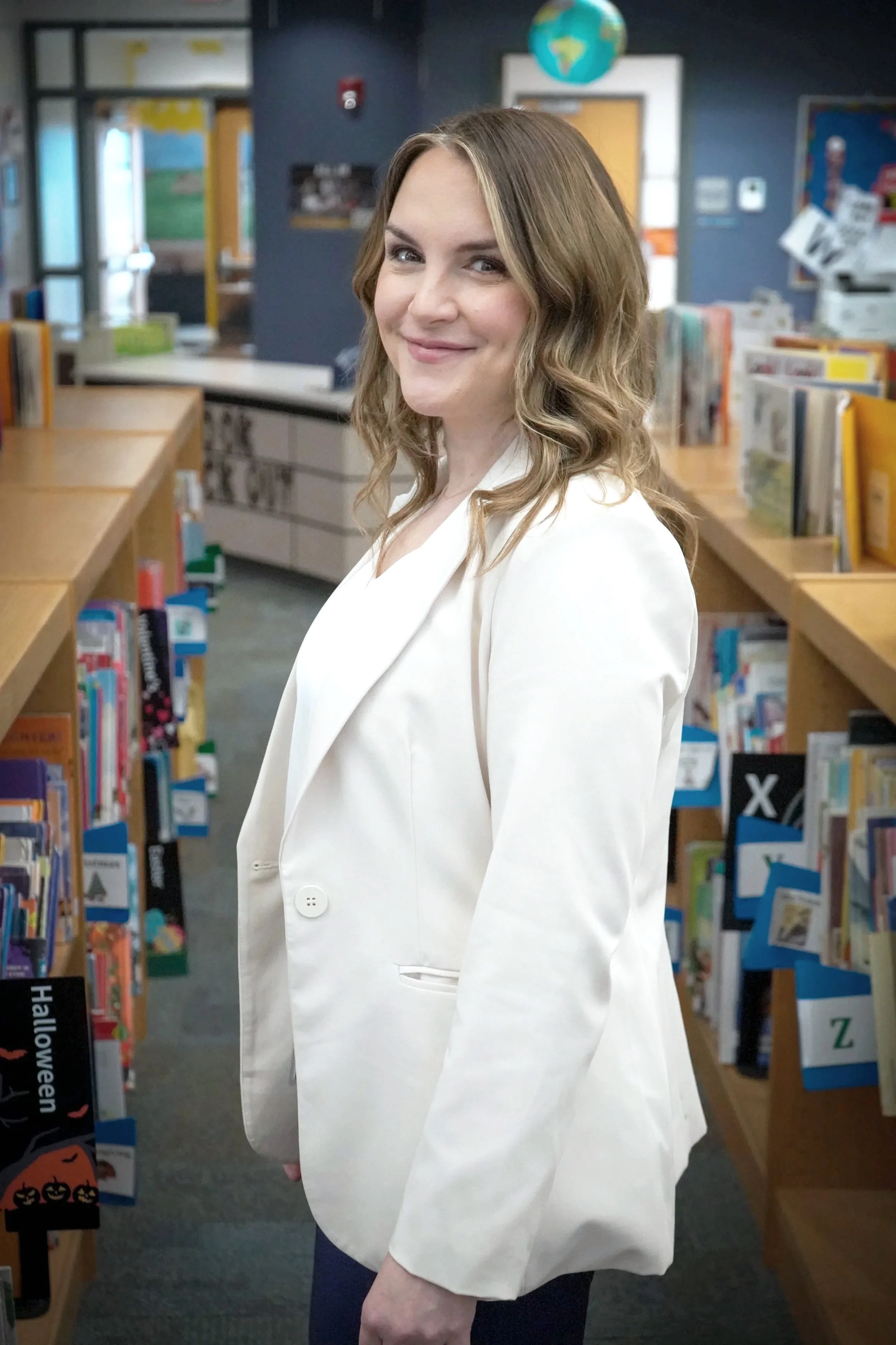 A woman with wavy blonde hair and a white blazer smiling in a library with bookshelves and a world globe in the background.
