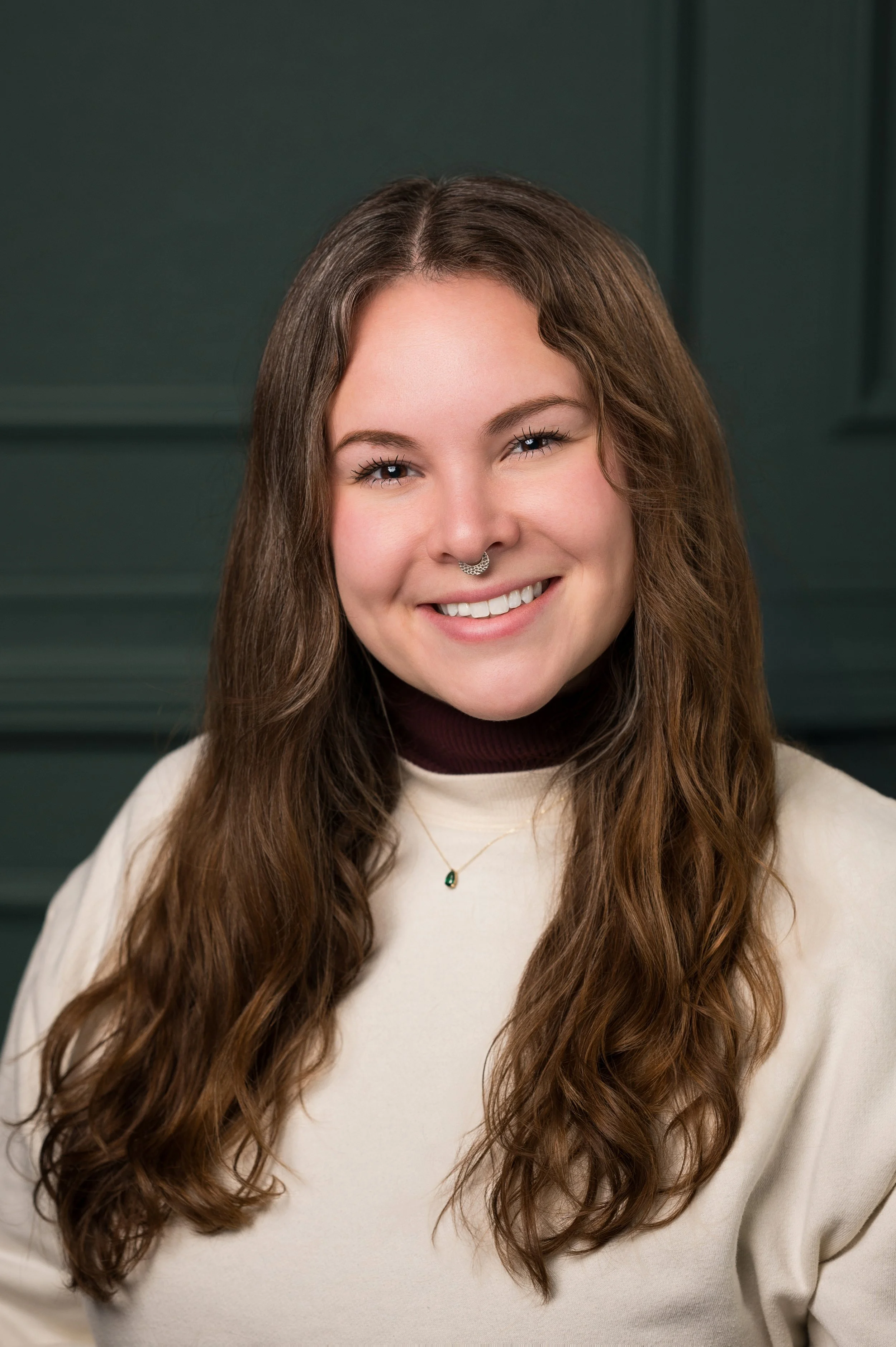 Close-up of a young woman with long wavy brown hair smiling, wearing a beige sweater, a dark turtleneck underneath, a delicate necklace with a green pendant, and a nose piercing, standing against a dark green paneled background.