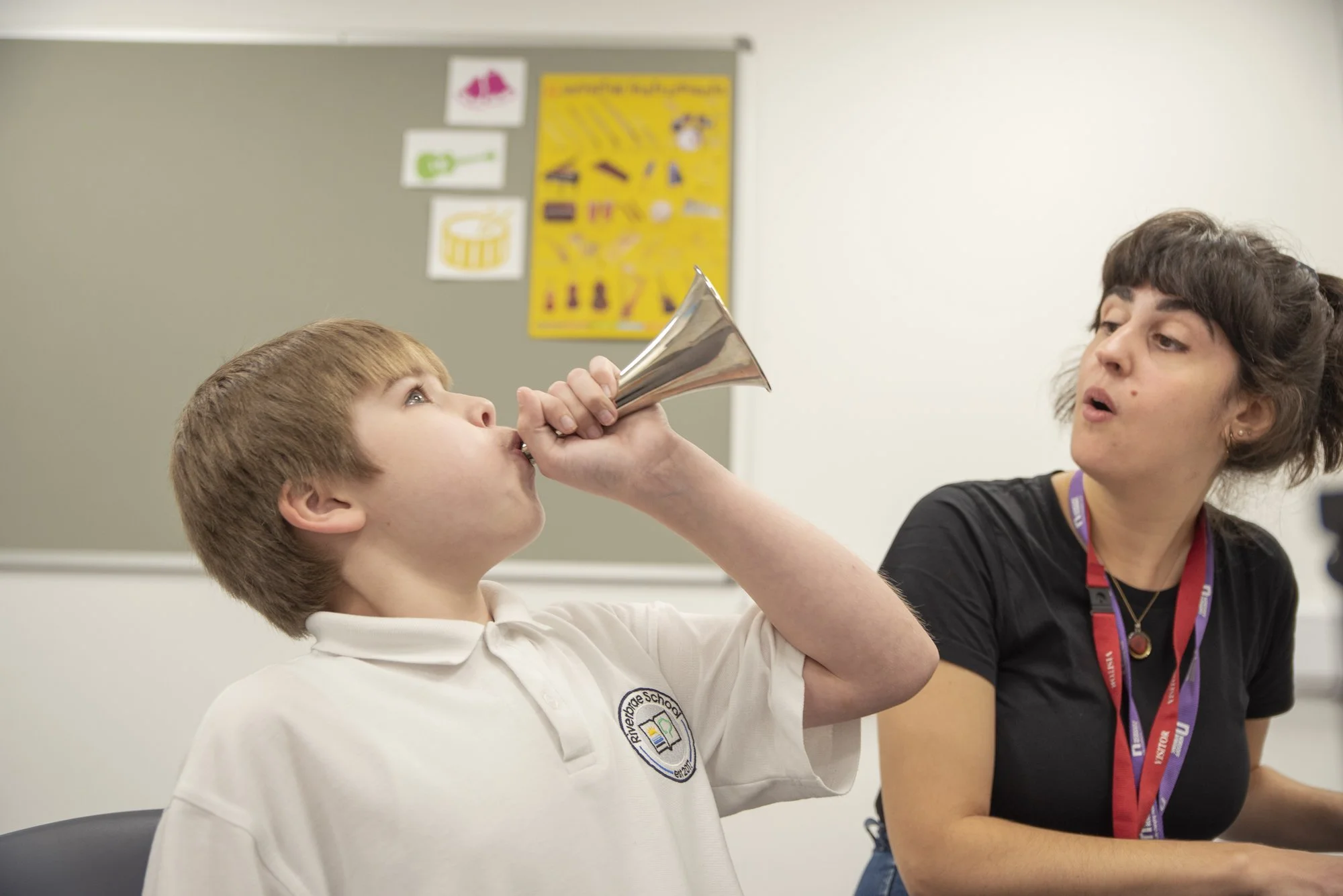 A young boy in a school uniform plays a small silver horn whilst accompanied by a woman at the piano.