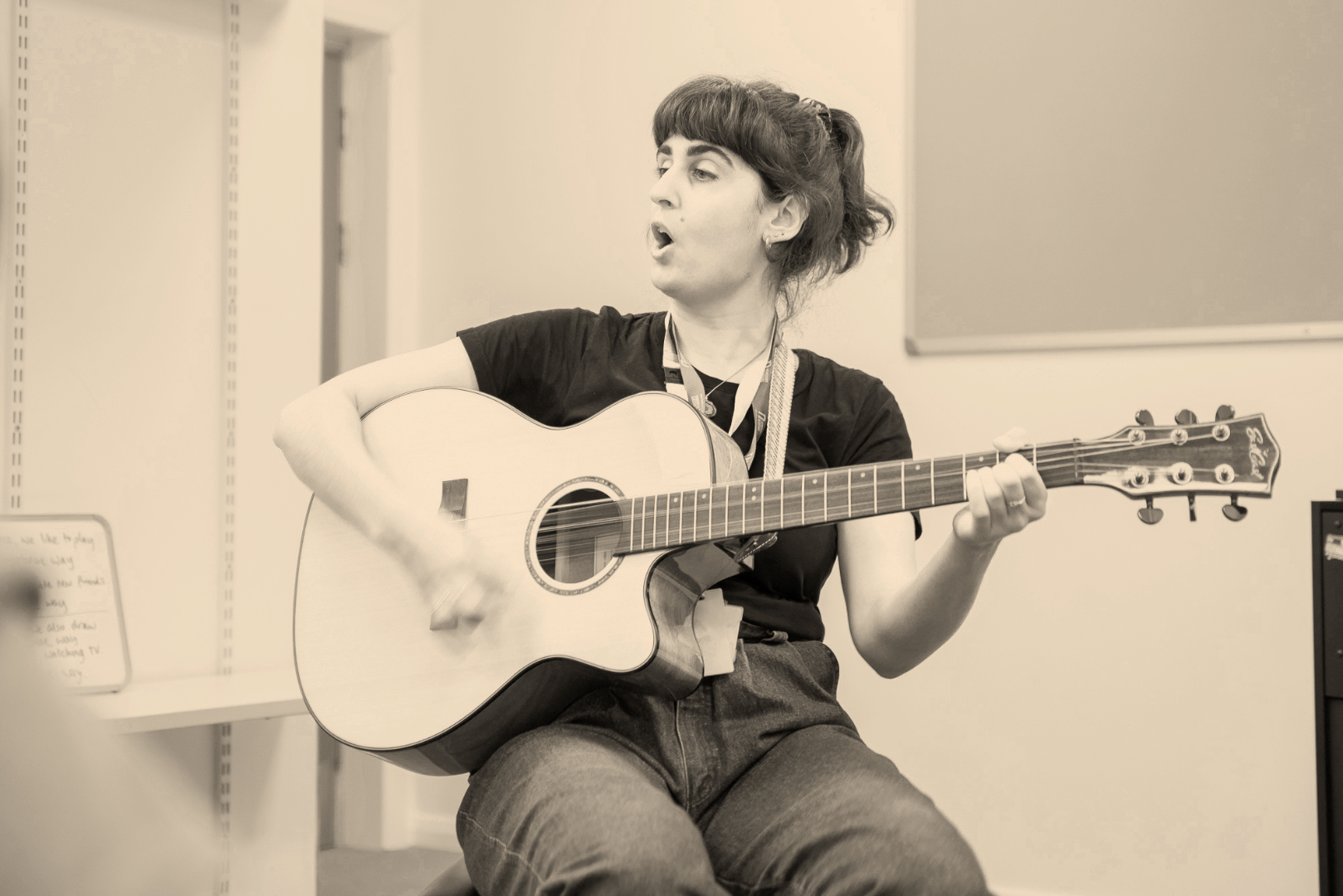 A woman playing an acoustic guitar and singing, seated in a room.