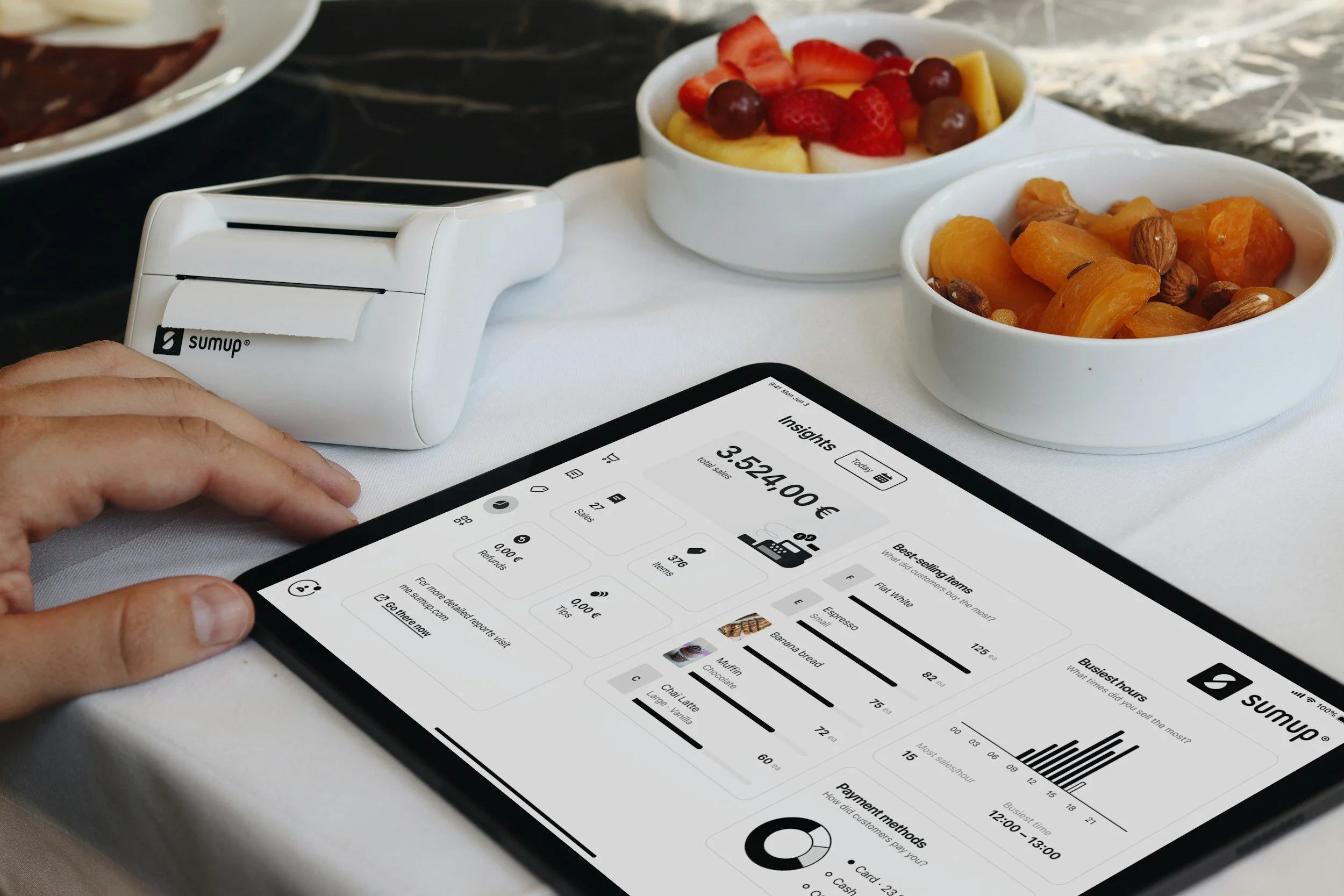 A person using a SumUp card reader to make a payment with a tablet displaying a sales report, with bowls of fruit and nuts on the table.