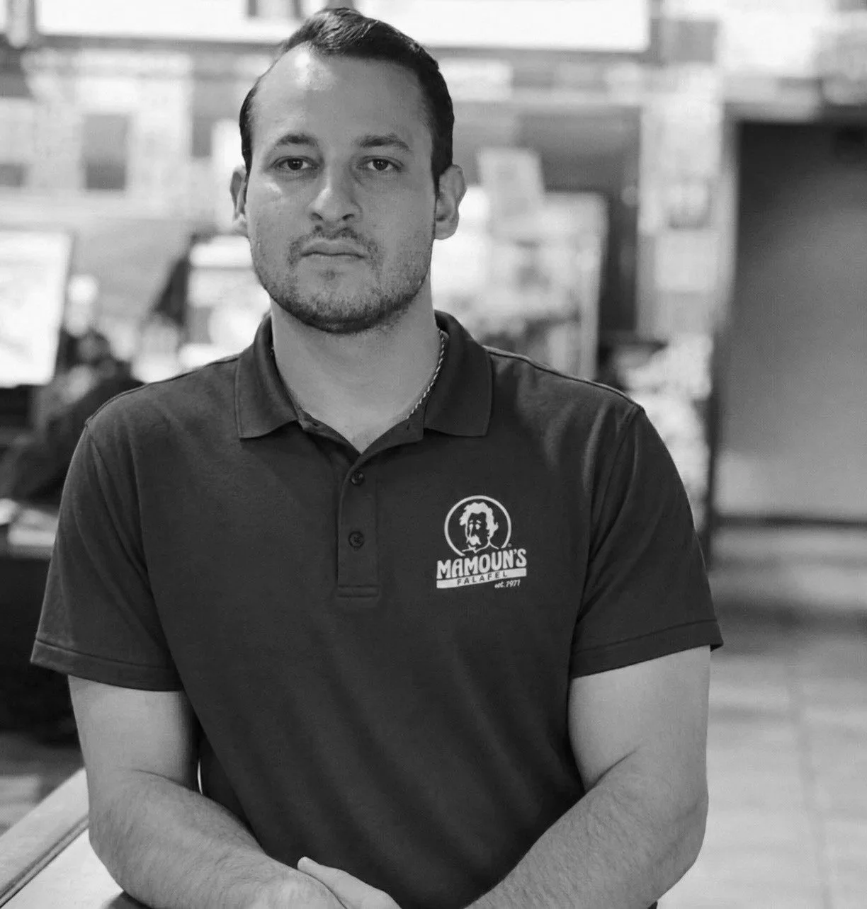 A man with short, dark hair and a goatee, wearing a black polo shirt with a logo that says Mamoun's Falafel, standing in a restaurant with a blurred background.