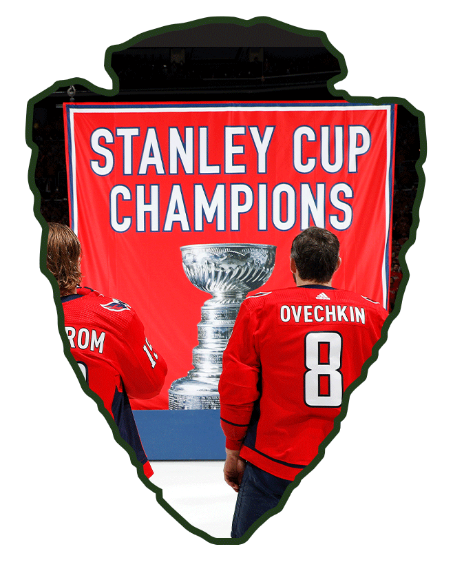 Two hockey players in red jerseys stand in front of a banner that reads "Stanley Cup Champions" with an image of the Stanley Cup trophy.