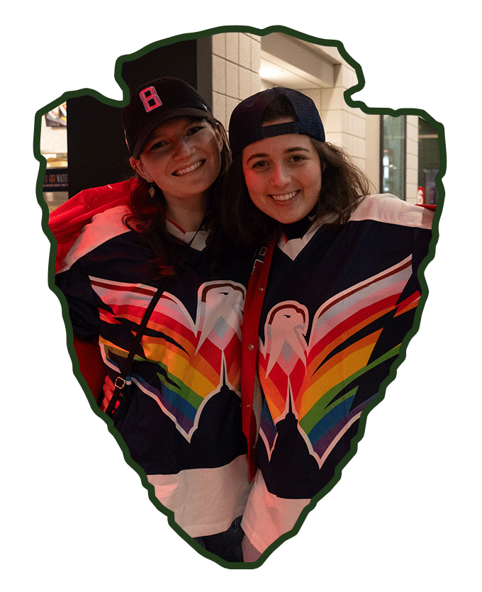 Two women smiling and wearing rainbow-themed sports jerseys, standing close together inside a heart-shaped outline.