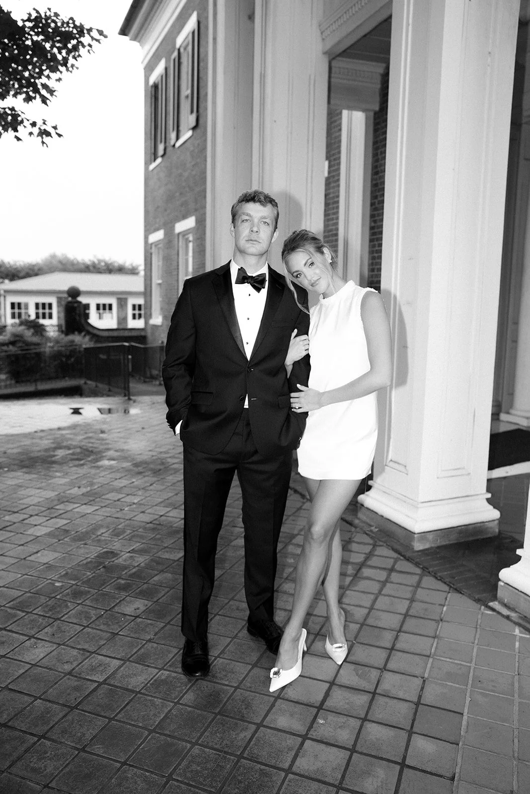 A man in a tuxedo and a woman in a white dress and high heels pose together outside a building in black and white.
