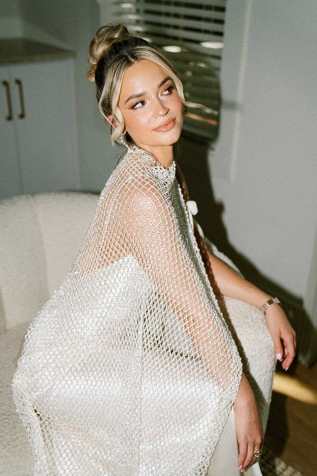 A woman with blonde hair styled in a high bun, wearing a beaded, netted cape over a white dress, sitting on a chair, looking to the side in a modern room.